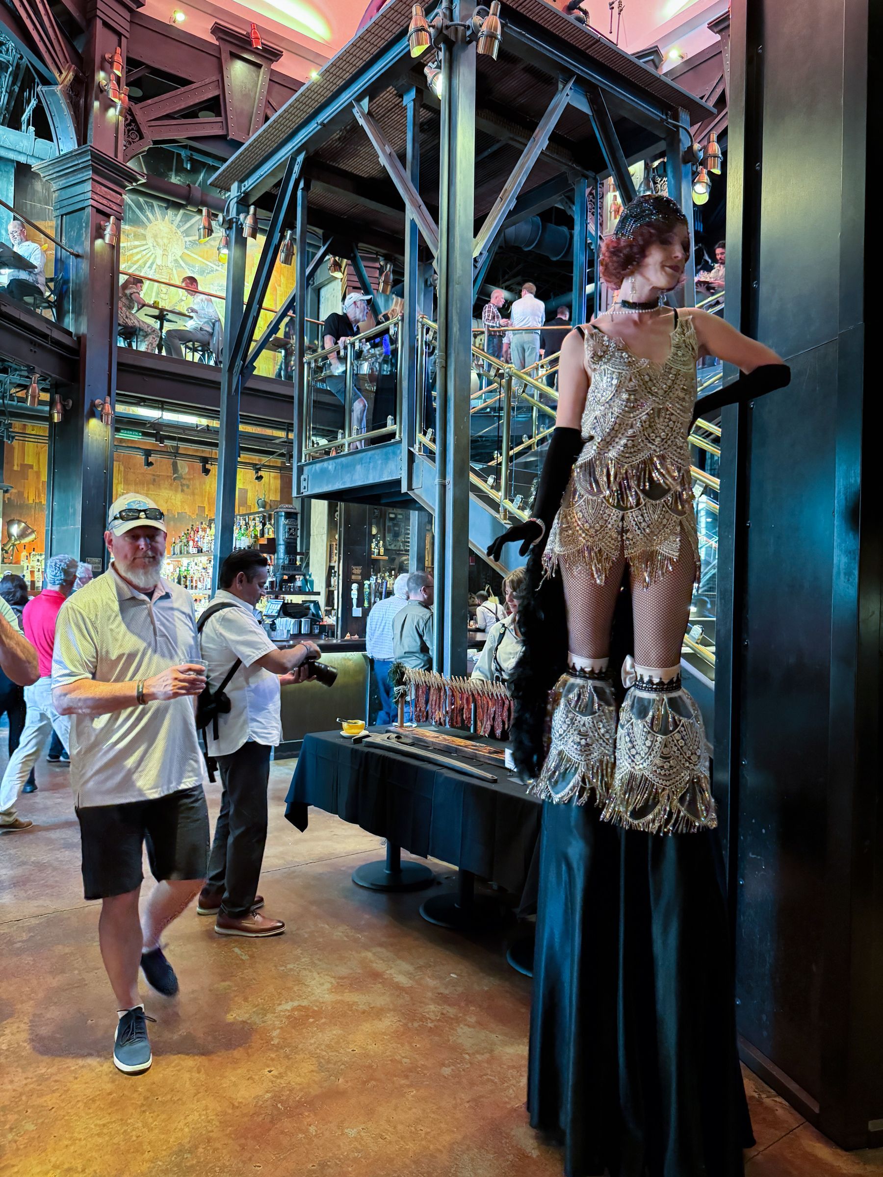 Woman on stilts in gold outfit, posing indoors. People nearby, decorative industrial setting.