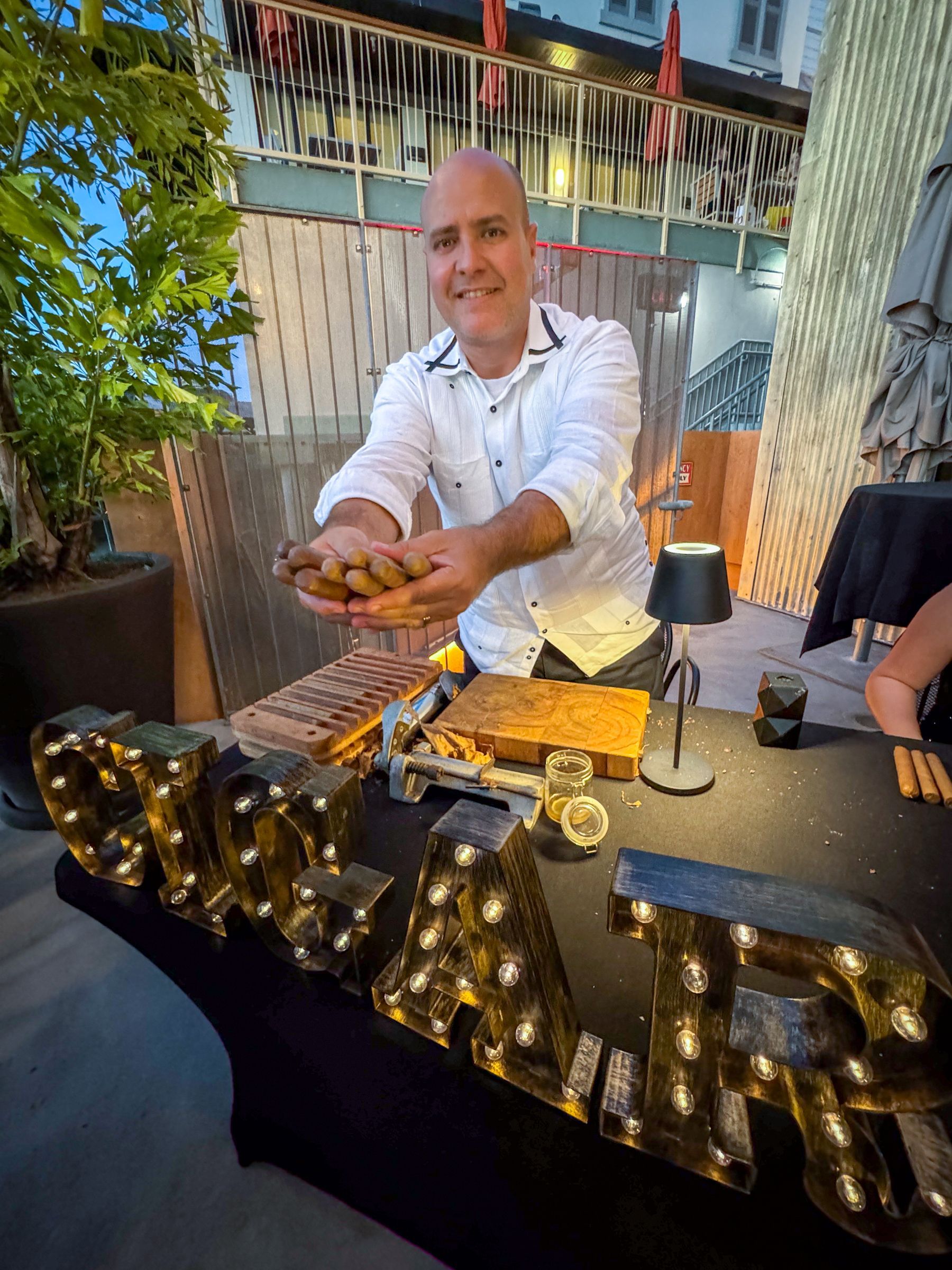 Man holding cigars, standing behind a table with lit sign and a lamp. Outdoor setting.