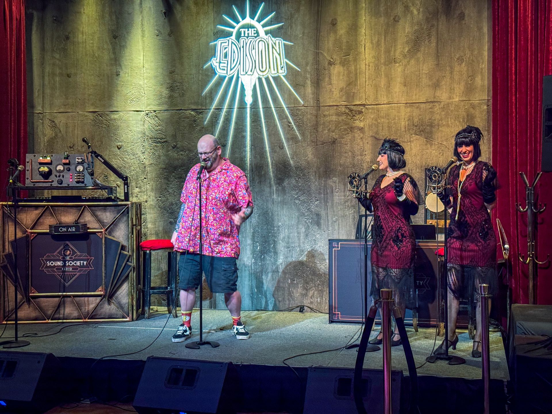 A man in pink shirt and two women in flapper dresses on stage; a band performing in front of a logo.