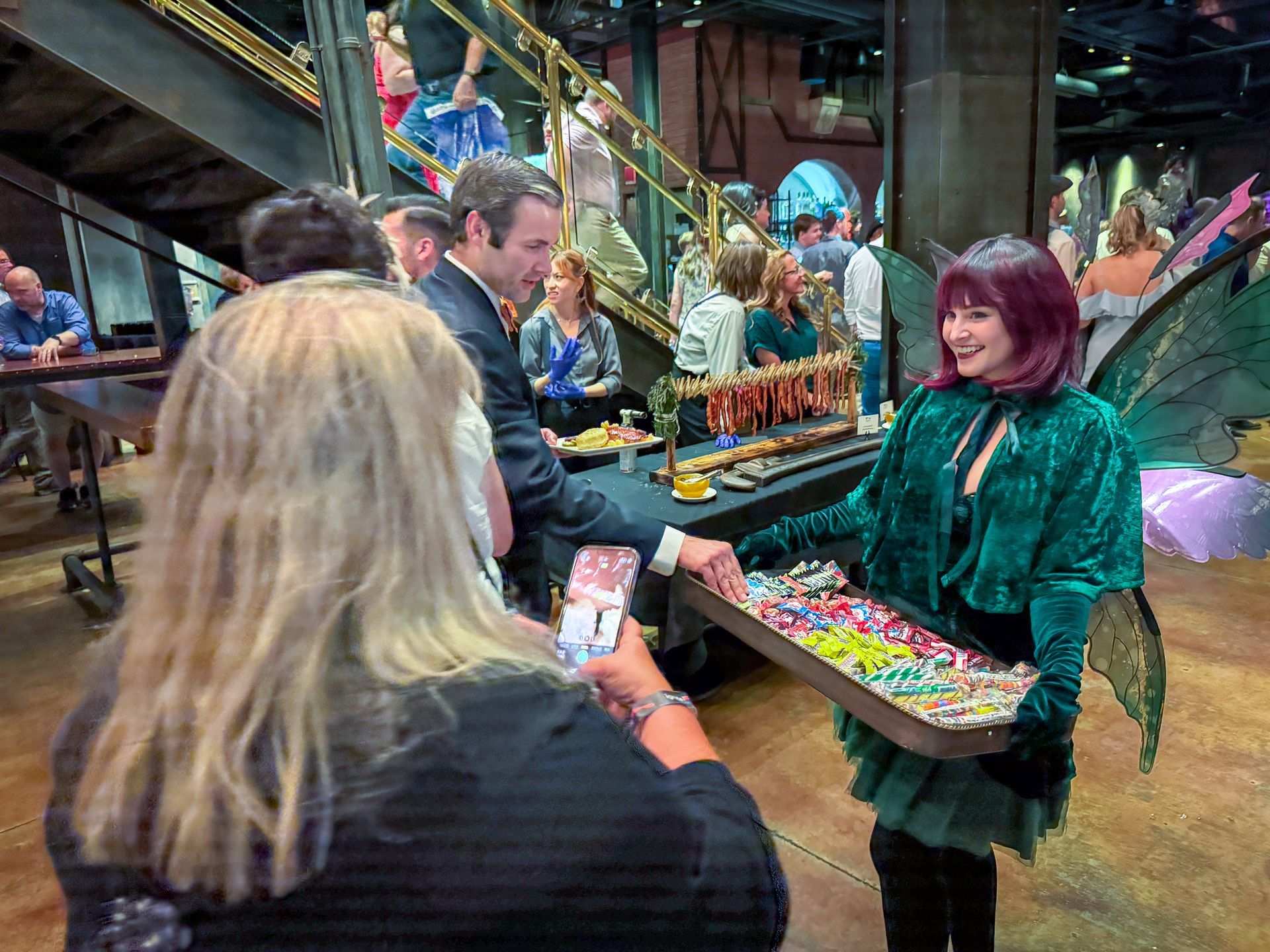Woman in fairy costume offers jewelry at event. Others are present at a buffet table, next to a staircase.