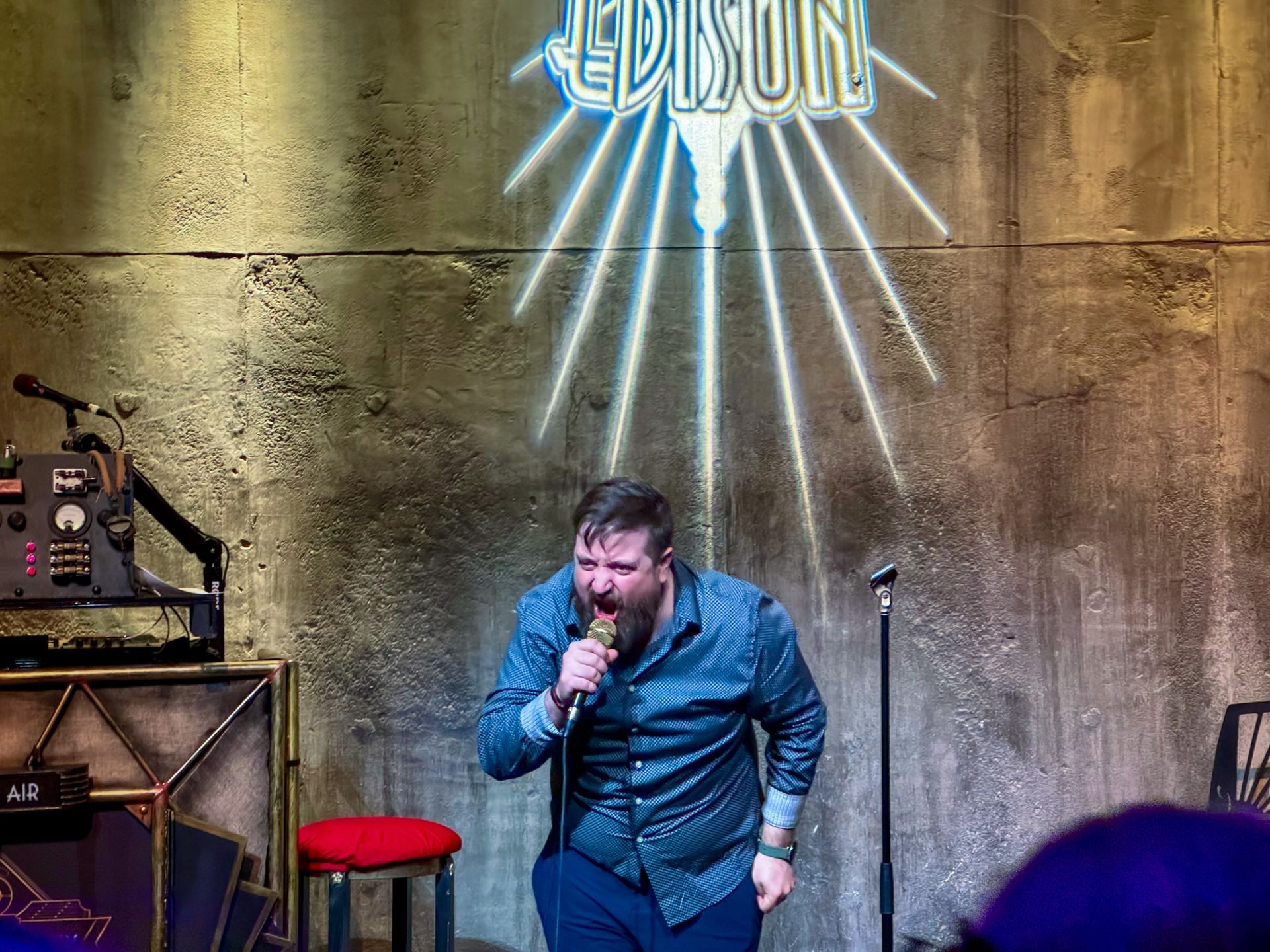 Man with microphone performing stand-up comedy at Edison venue; blue neon sign, red stool, concrete background.