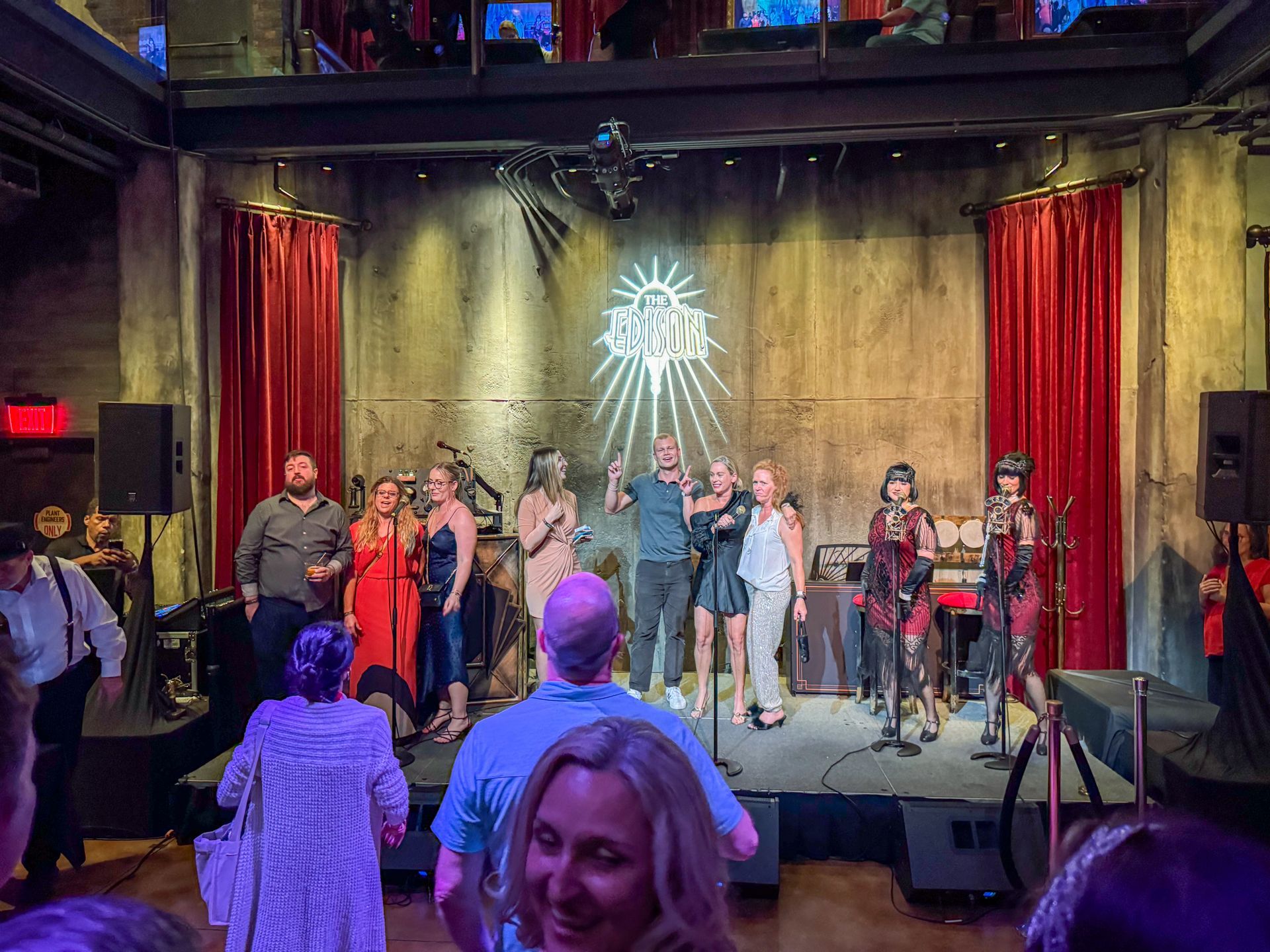 People on stage with microphones, lit by a sunburst. Red curtains frame the stage.