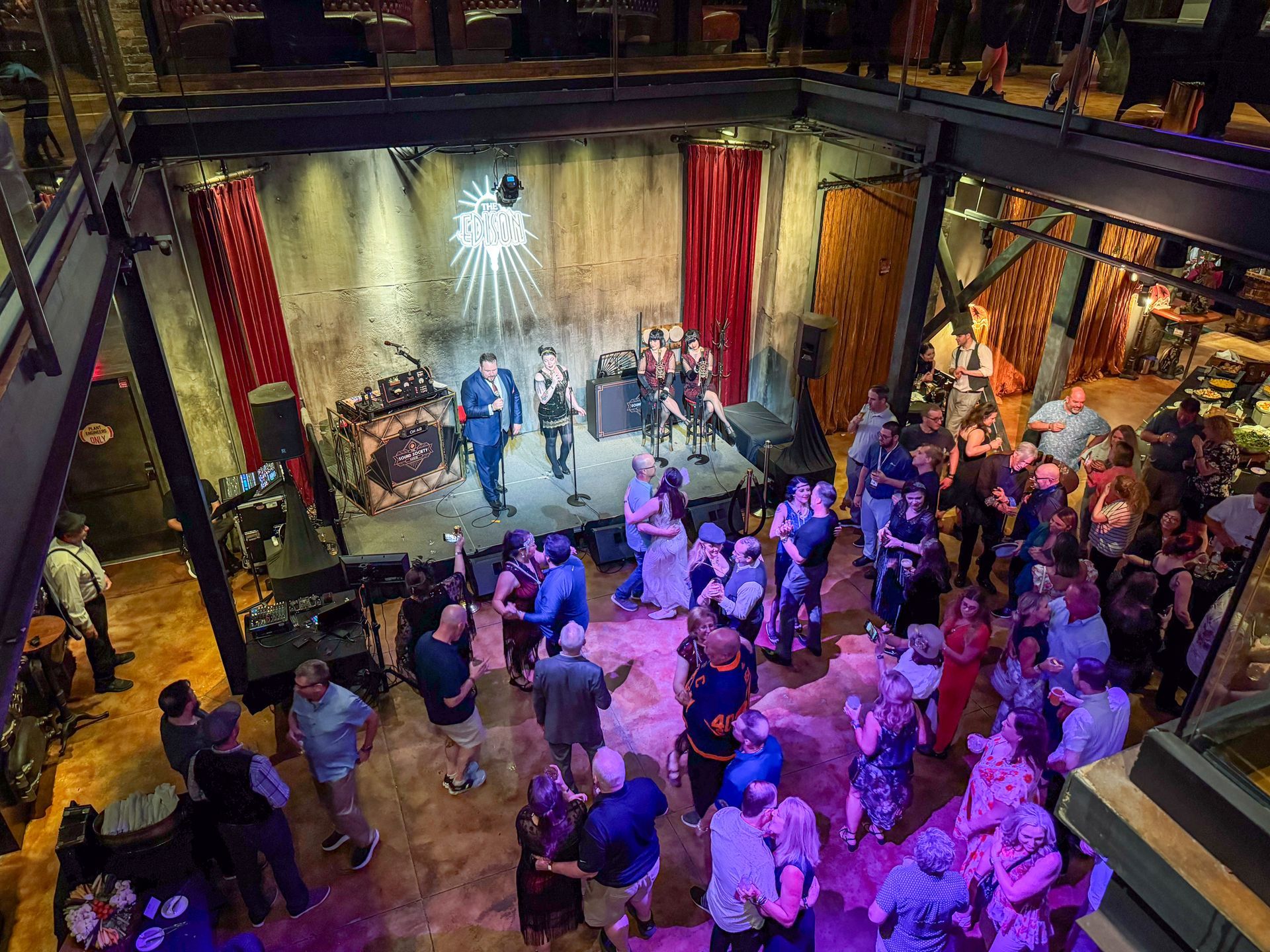 Band performing on stage in a dimly lit venue with a crowd dancing below. Red curtains and stone walls.