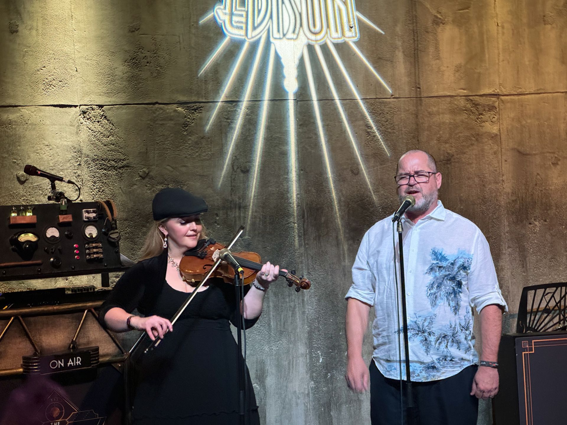 Woman plays violin, man sings at Edison club. Stage with art deco decor.