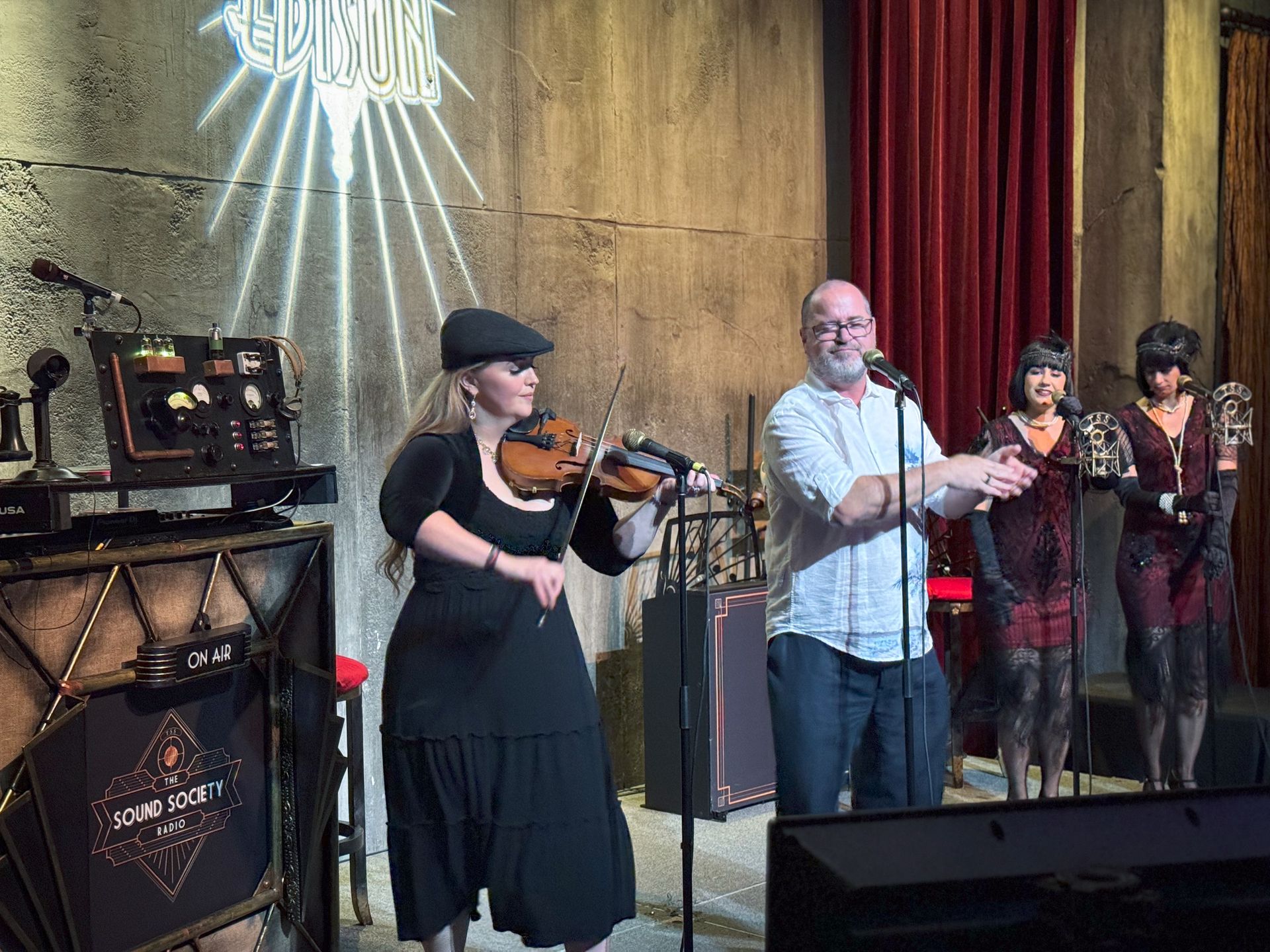 Musicians perform on stage: violinist, singer, and two women in flapper dresses. The setting is dimly lit.