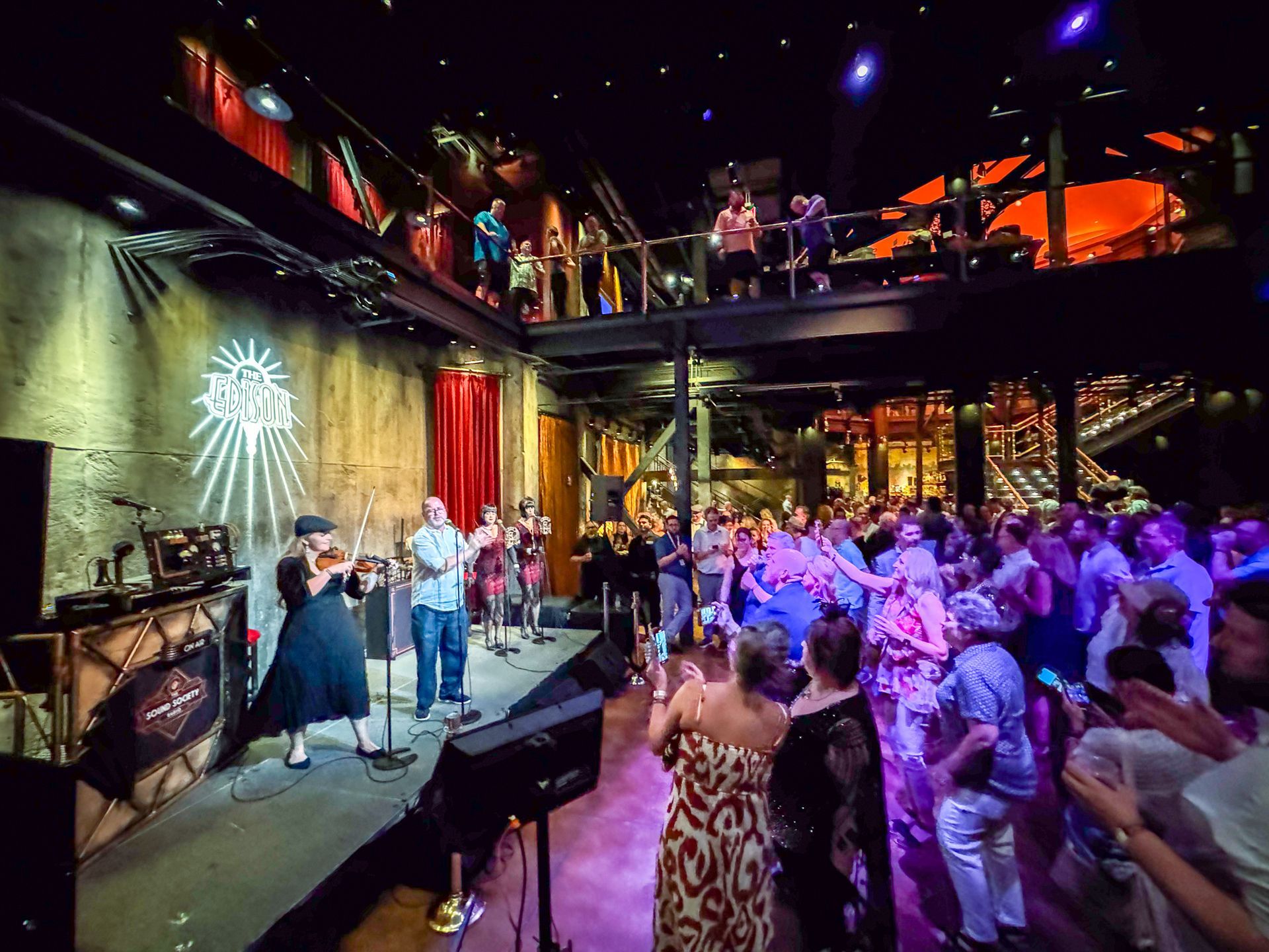 Band performs on stage in a rustic venue with a crowd clapping and watching. Balcony above.