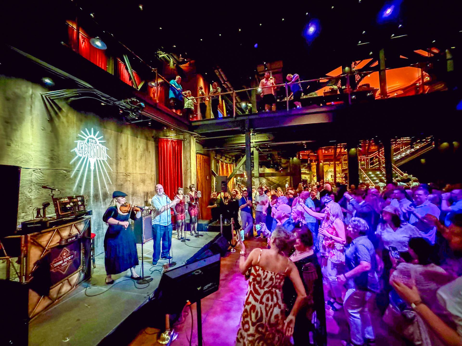Band performing on stage in a crowded music venue. People dance and watch from a balcony.