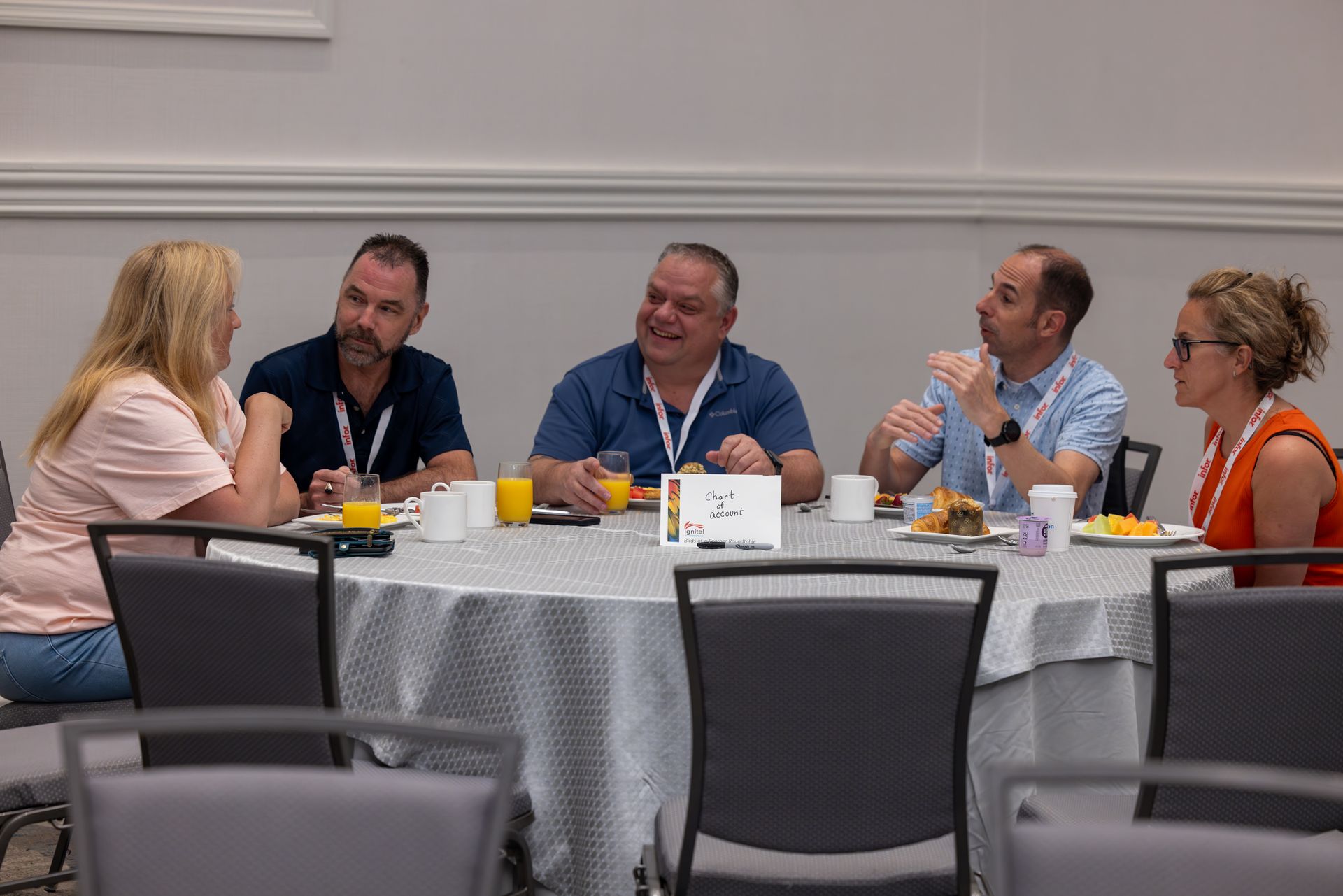Five people at a round table, eating breakfast and talking. The setting is a conference room.