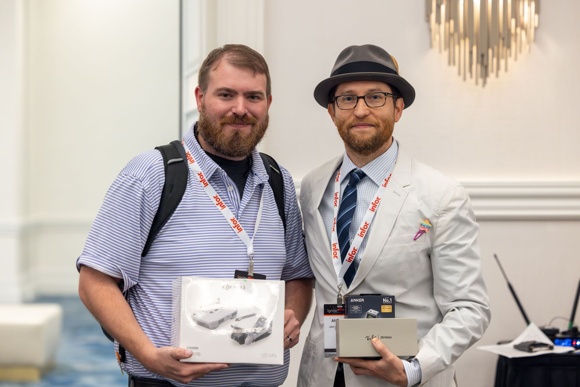 Two men holding boxes, likely prizes, at an event. One wears a hat and tie, the other has a beard and backpack.