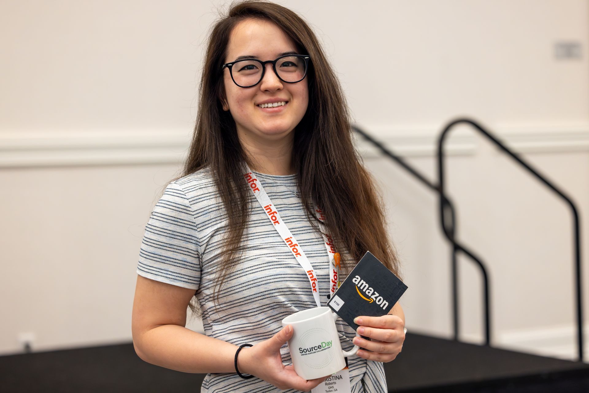 Woman with glasses smiles, holding a mug and a card, wearing a patterned dress. Standing indoors near steps.