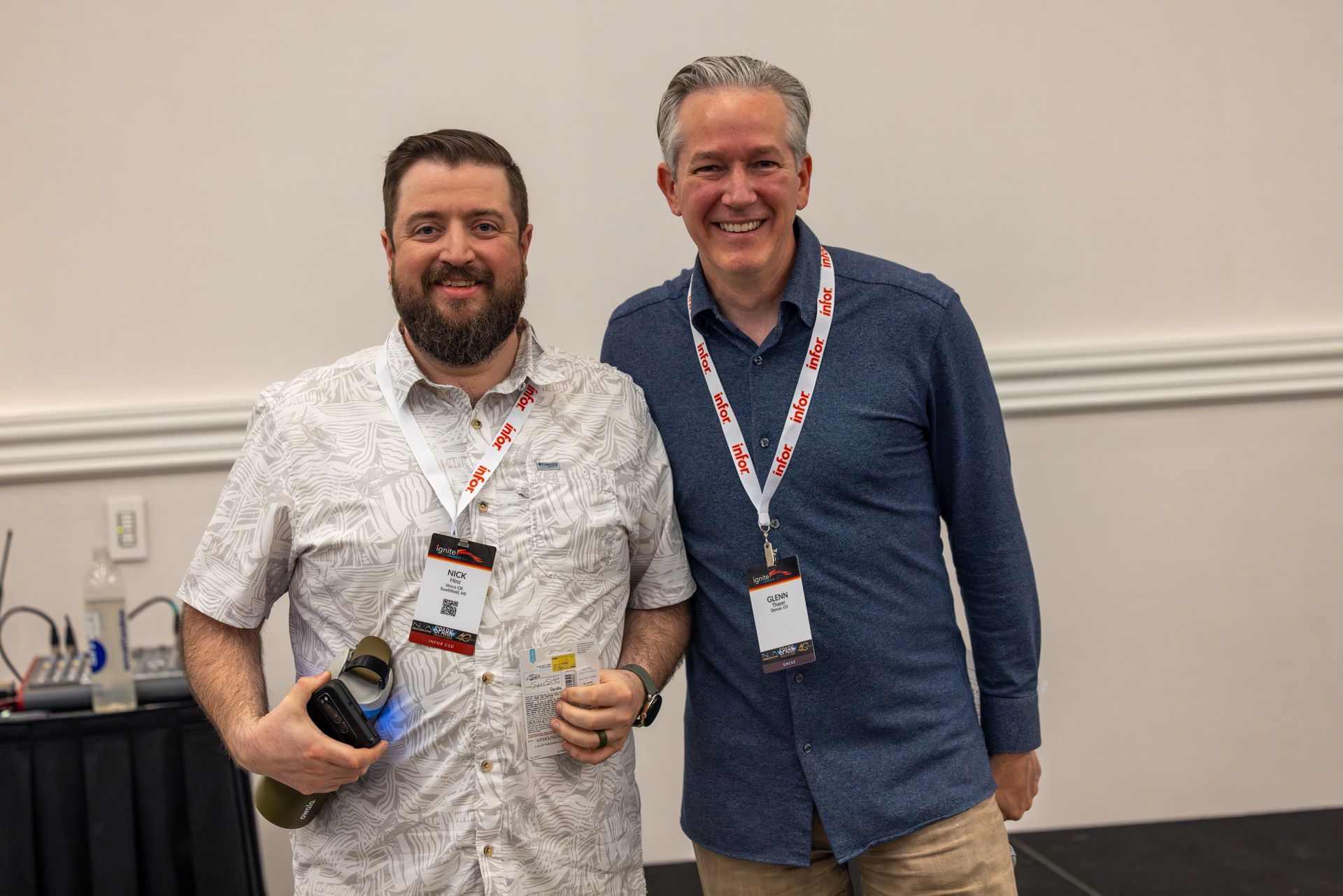 Two men smiling, wearing lanyards, standing next to each other. One man holds a bottle and a small item. 