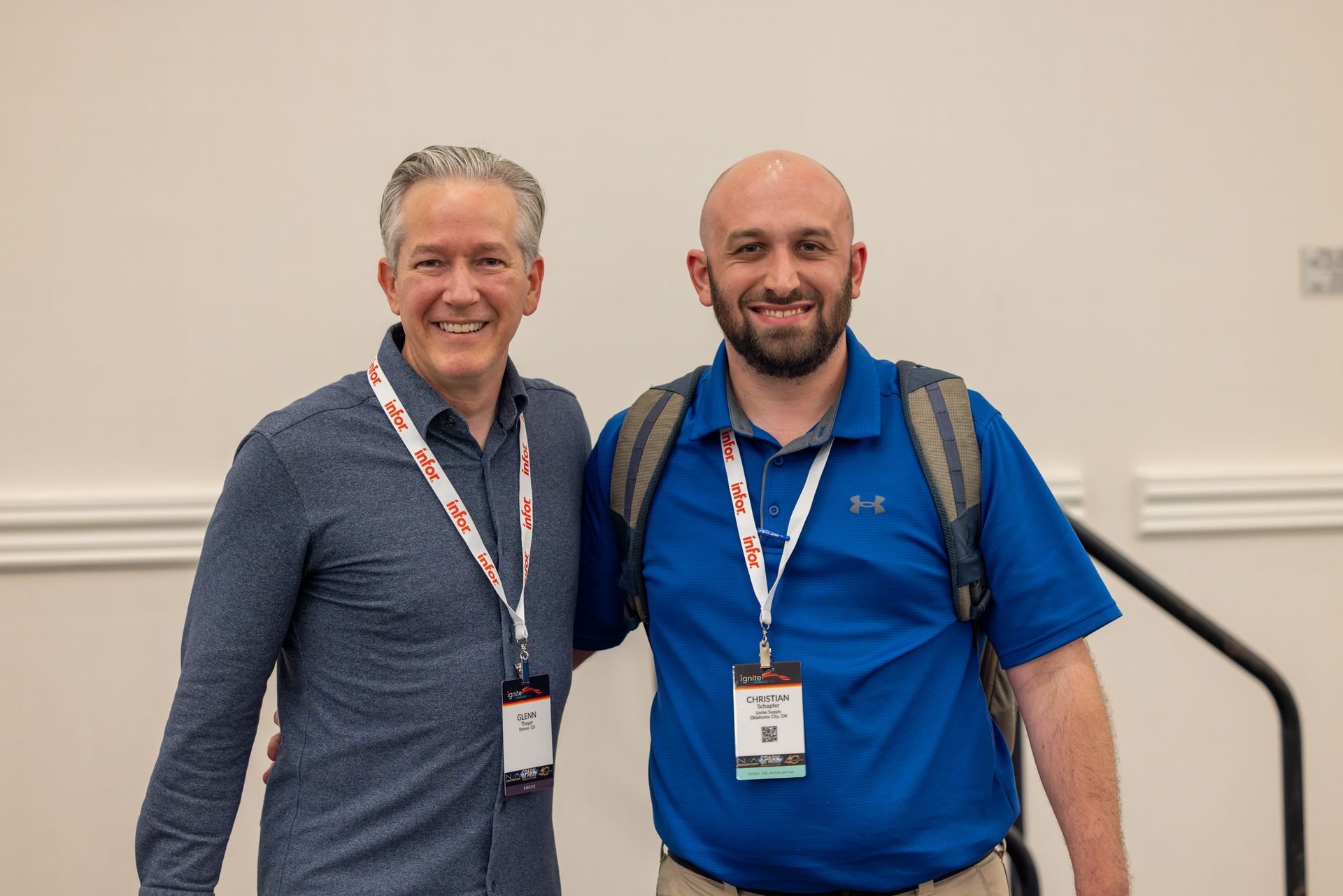 Two men posing together, smiling. One in a blue shirt with a backpack; the other in a patterned shirt. Both wearing lanyards.