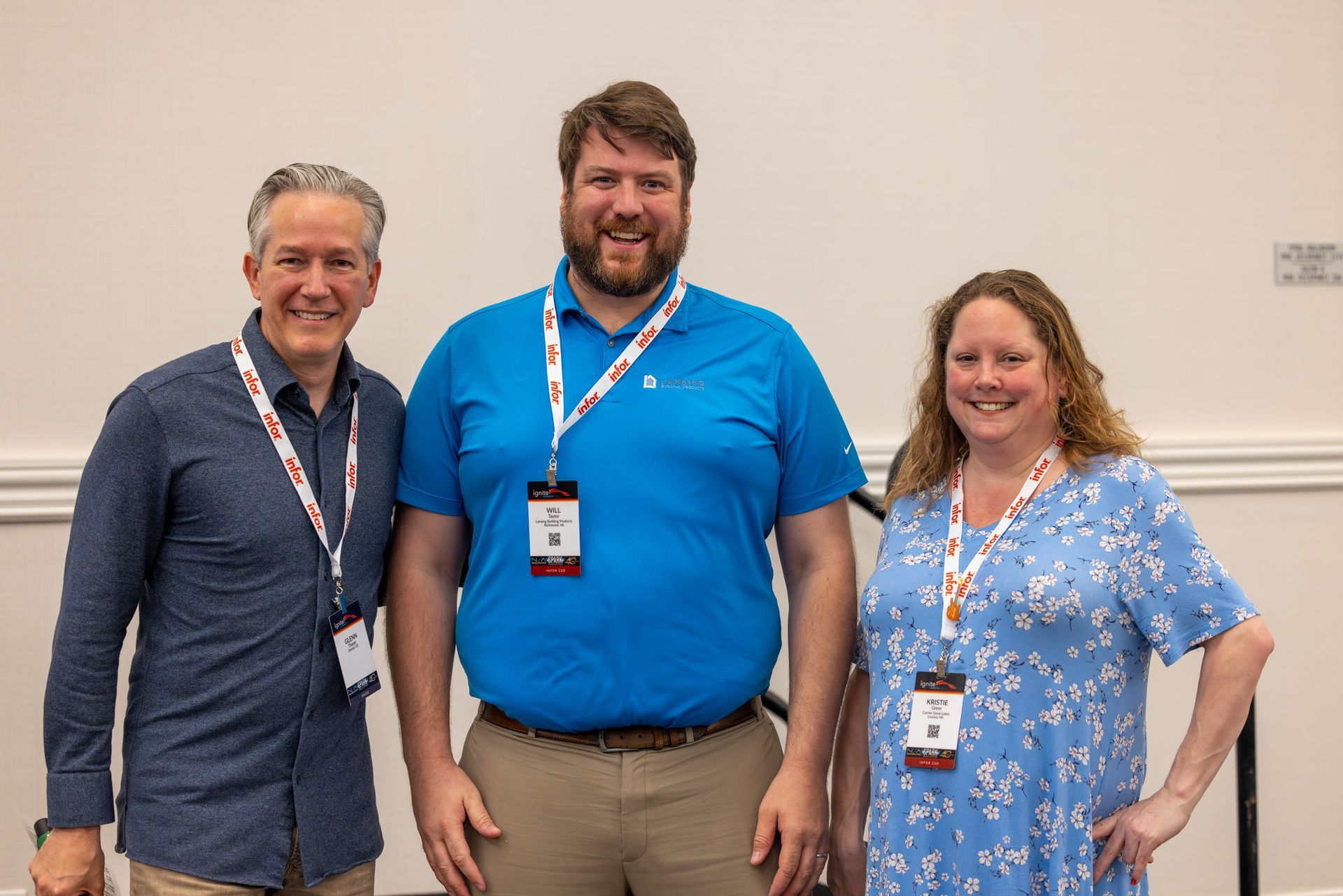 Three people smiling at a conference. The man in the middle wears blue; the others wear patterned shirts. All have lanyards.