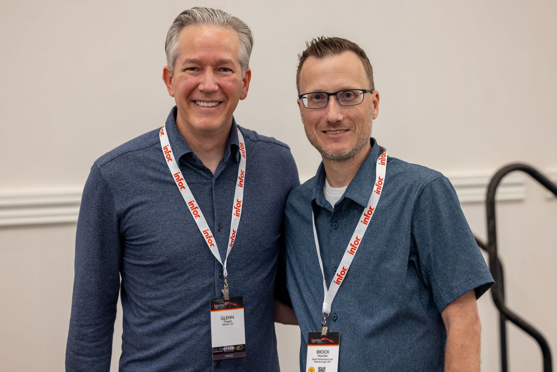 Two men with lanyards smile, posing close together. Indoors, against a light wall.