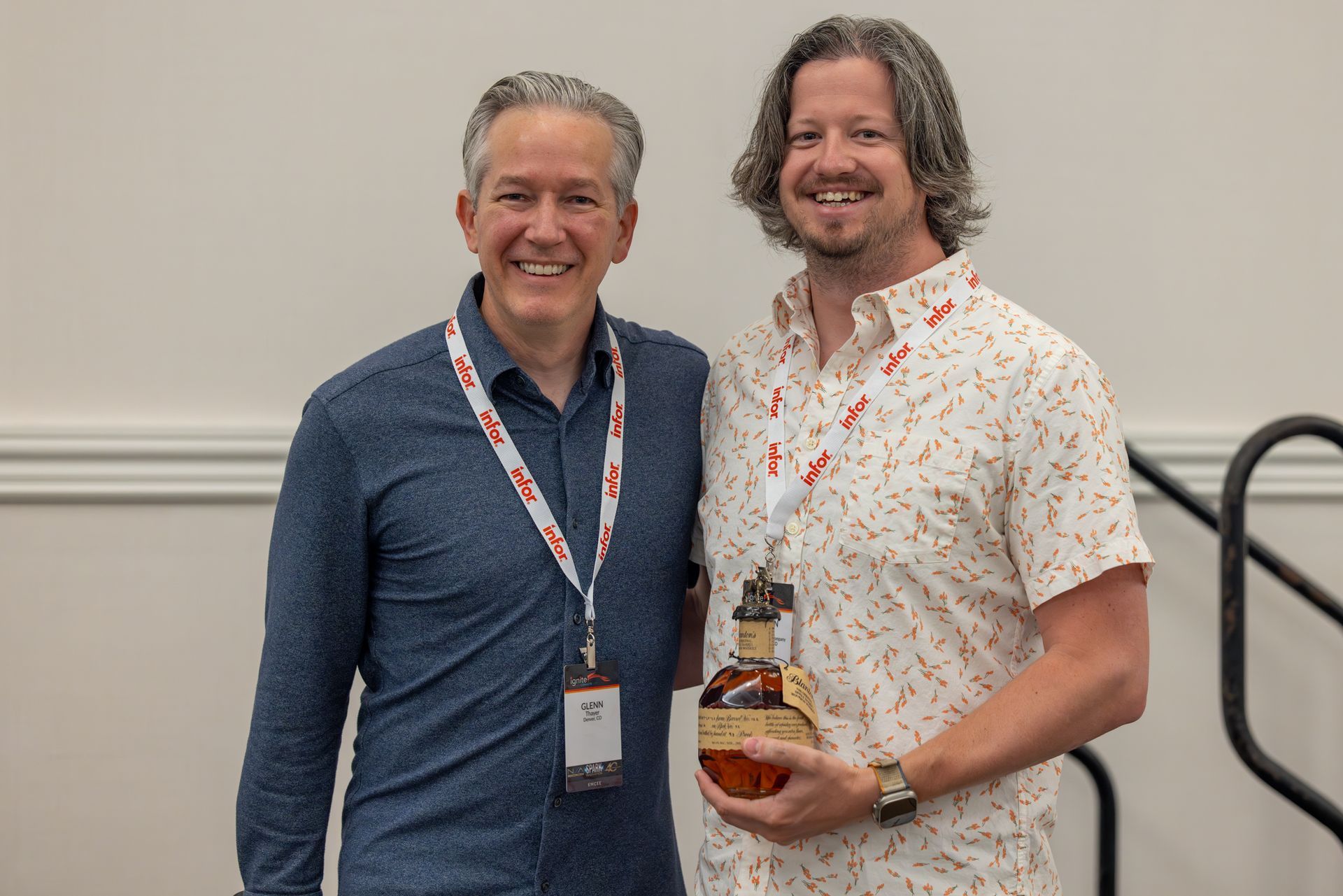 Two men smiling, one holding a whiskey bottle. Both wear lanyards, standing in a brightly lit room.