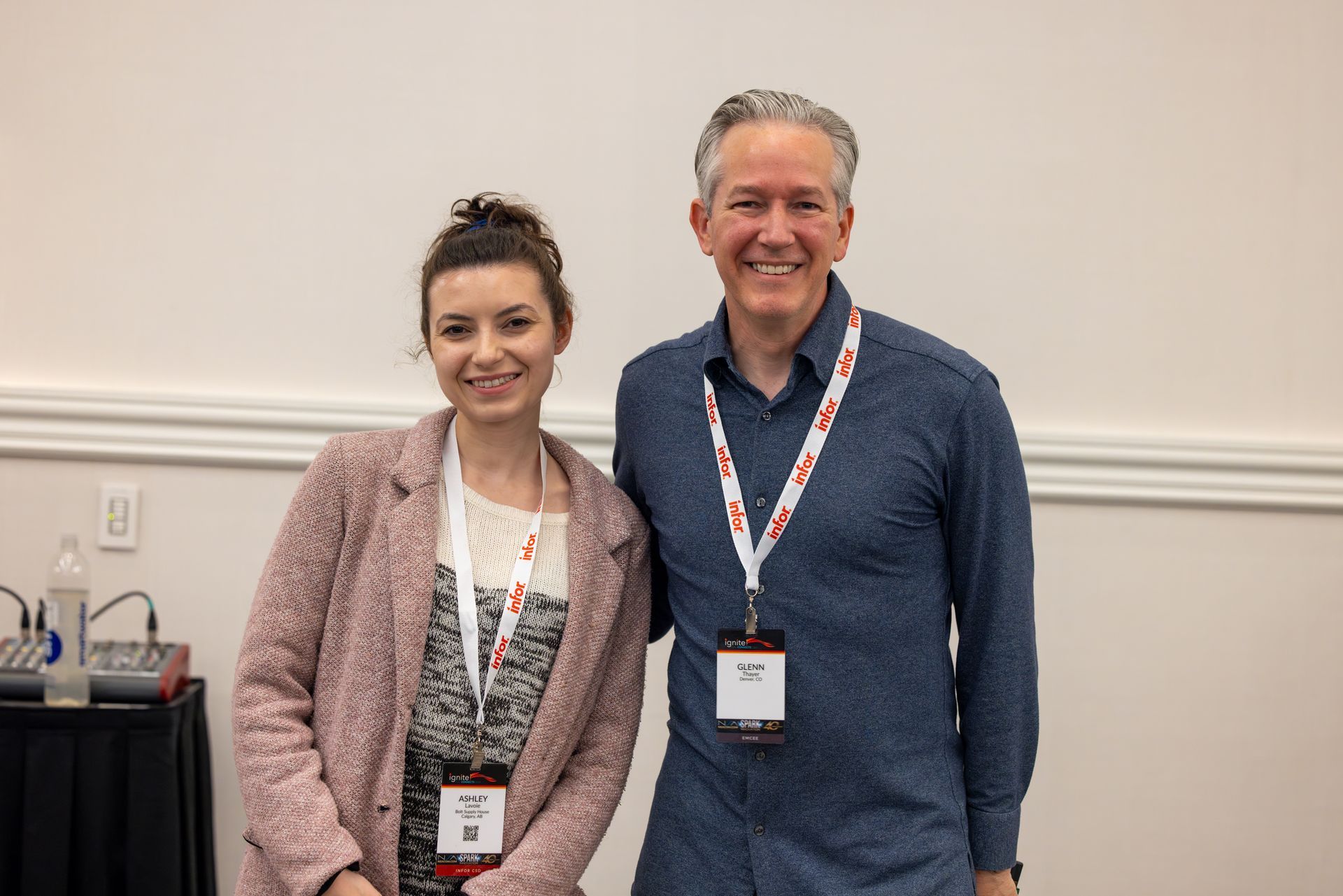 Two people posing together, both wearing lanyards, smiling in front of a white wall.
