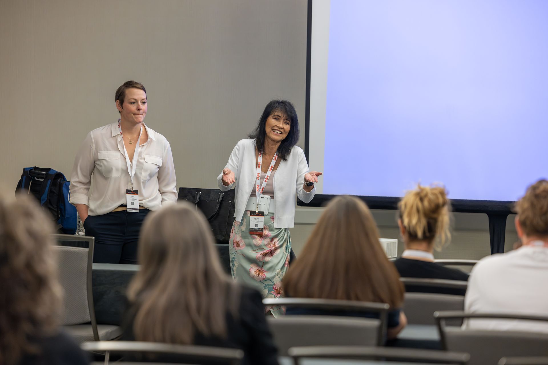 Two women present in a lecture hall. The presenter gestures, facing audience. A blank screen is behind them.