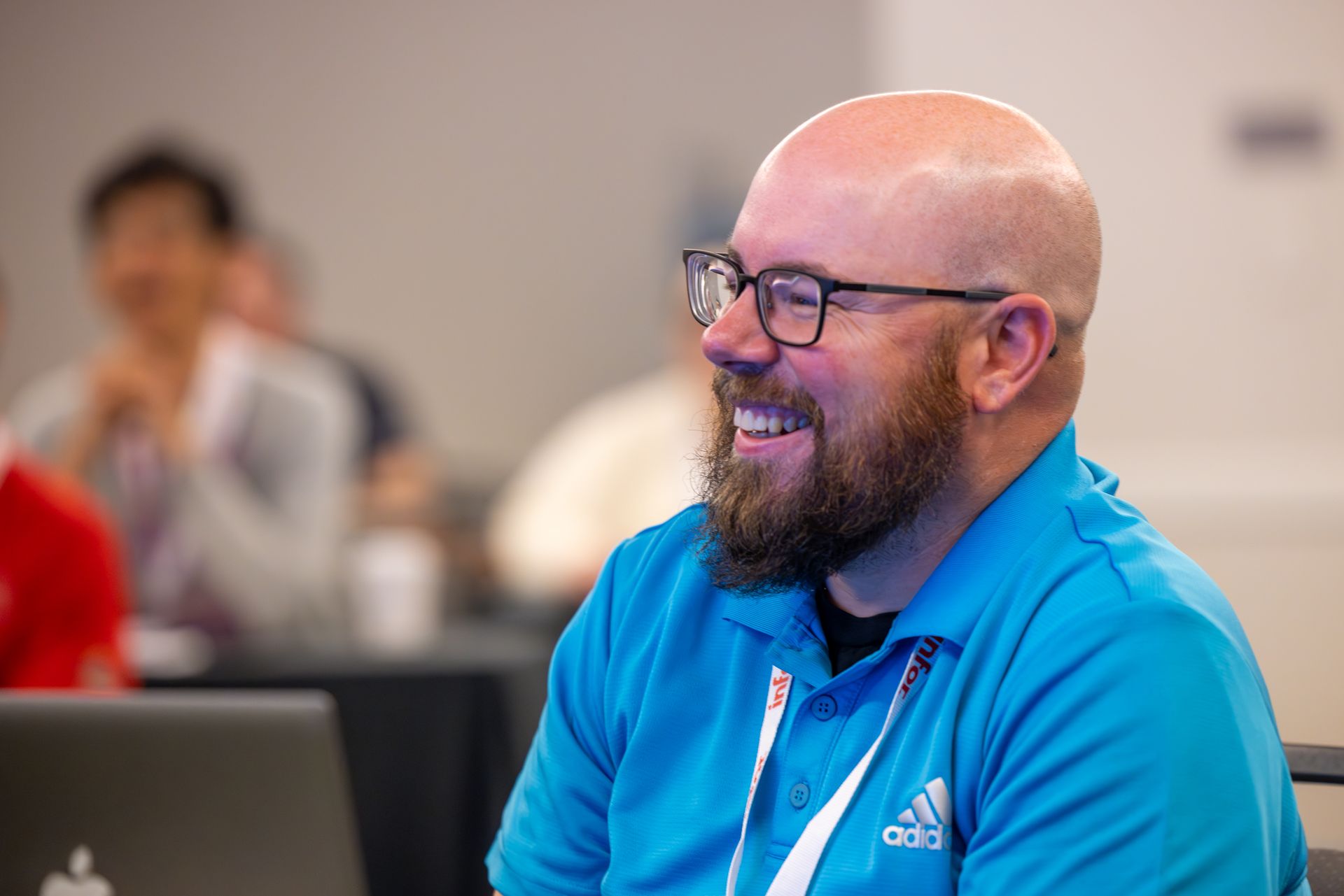 Man with glasses and beard smiles broadly, wearing a blue polo shirt, indoors.