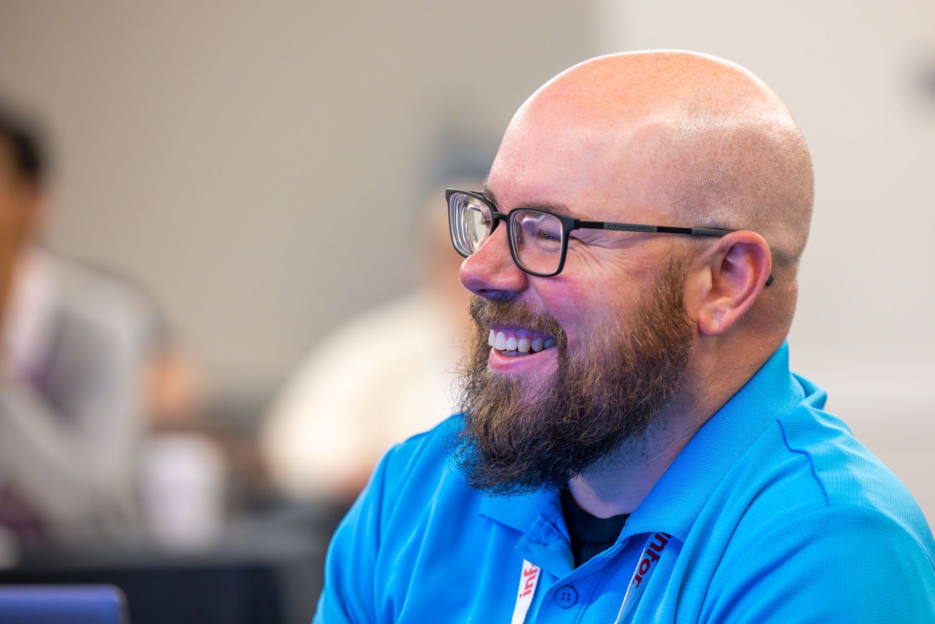 Bald man with glasses smiles widely, wearing a blue shirt, in a brightly lit room.