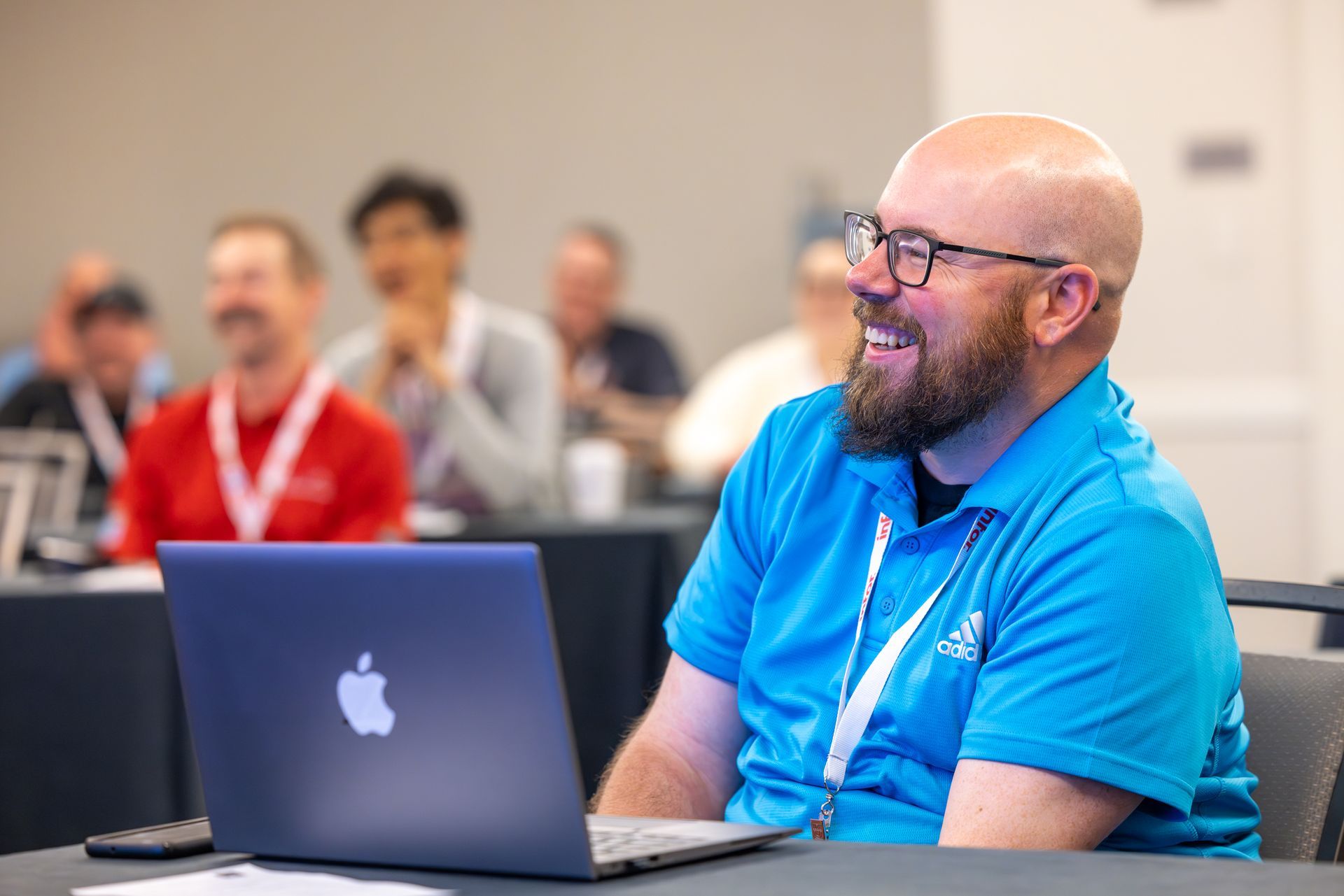Man in blue shirt smiles while using a laptop in a conference setting. Other attendees are in the background.