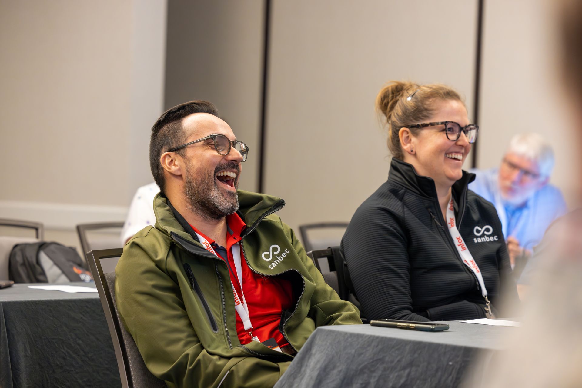 Two people laughing at a conference. Man in green jacket and woman in black jacket, both wearing glasses.