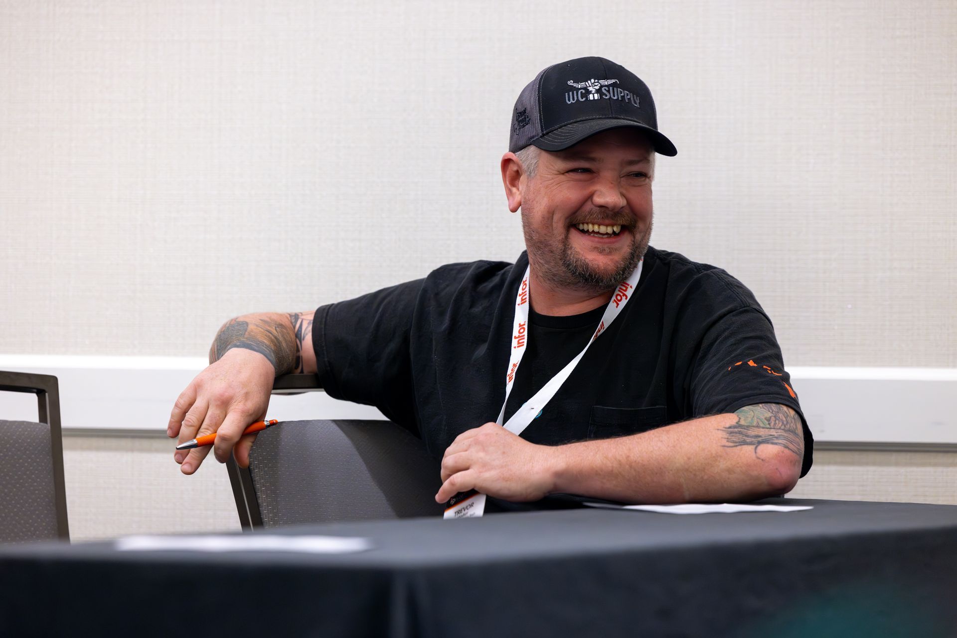 Man with tattoos smiles, wearing a black cap and shirt, sitting at a table with a name tag.