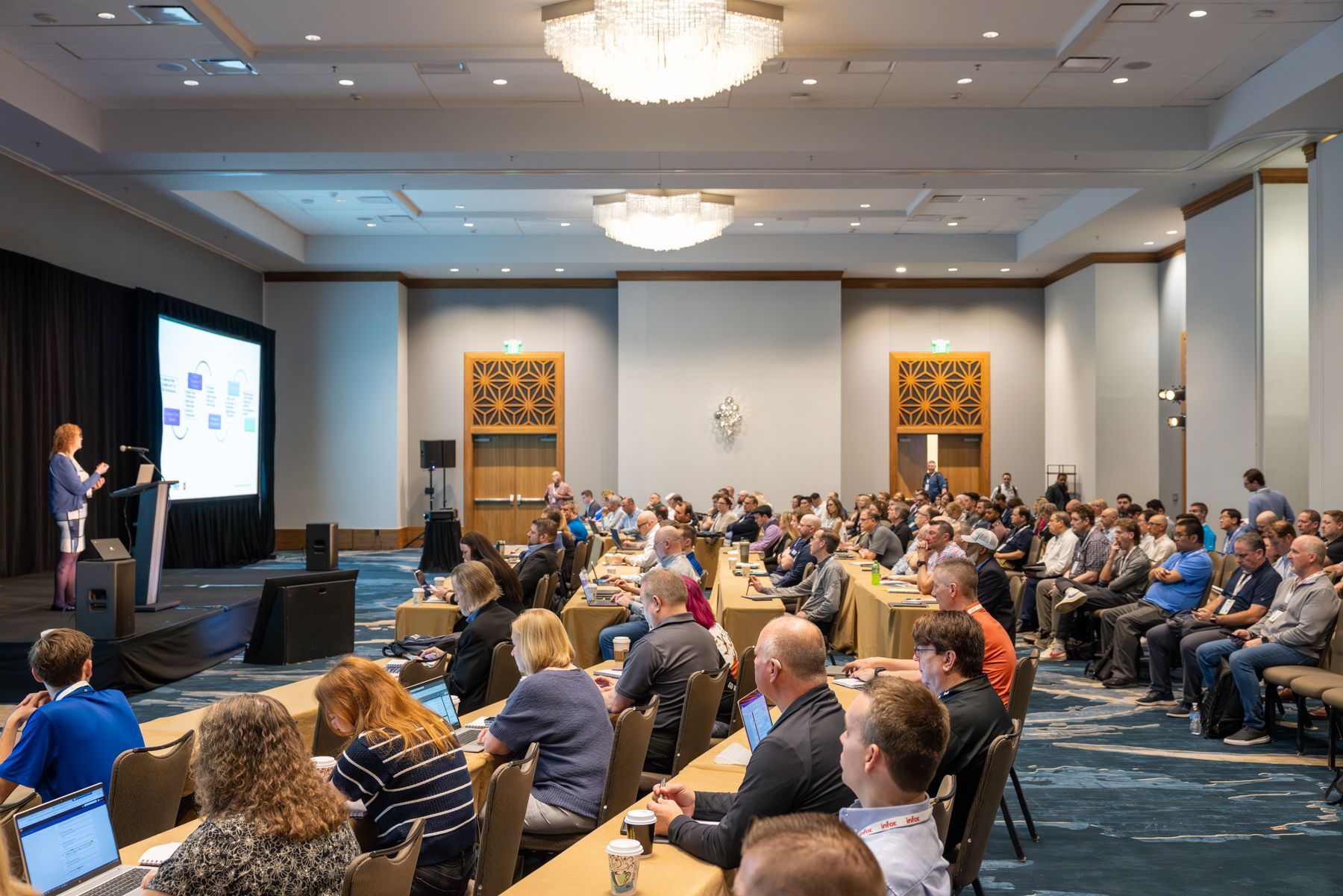 Woman speaking at a conference. Audience sits at tables facing the stage. A screen displays a presentation.