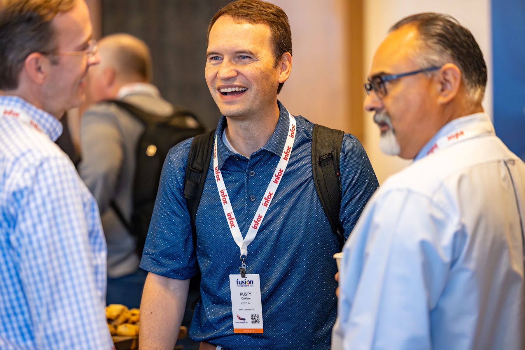 Three men in a conference setting, conversing. Man in blue shirt smiles, wearing a lanyard; other men wear glasses.