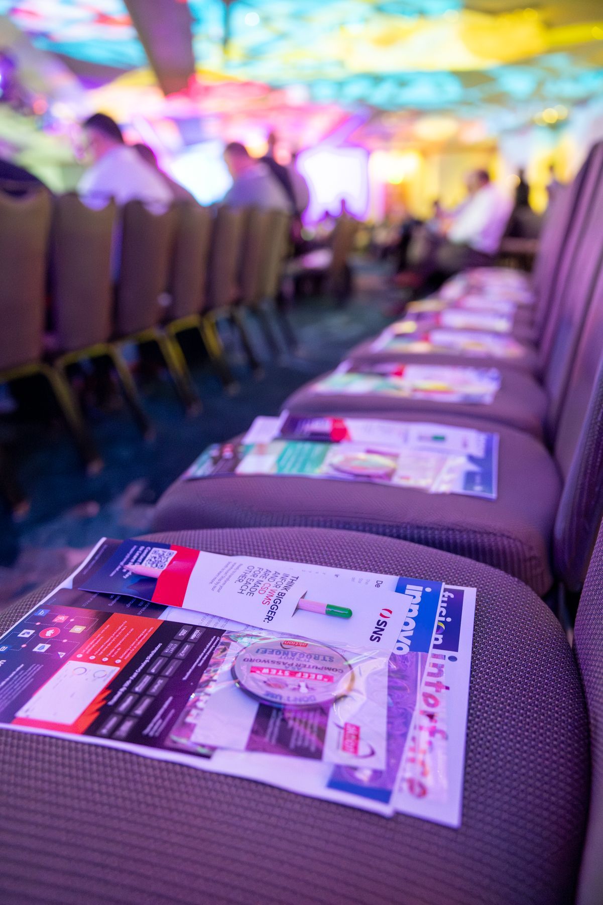 Row of conference chairs with handouts and a promotional item; event in progress, blurred background.