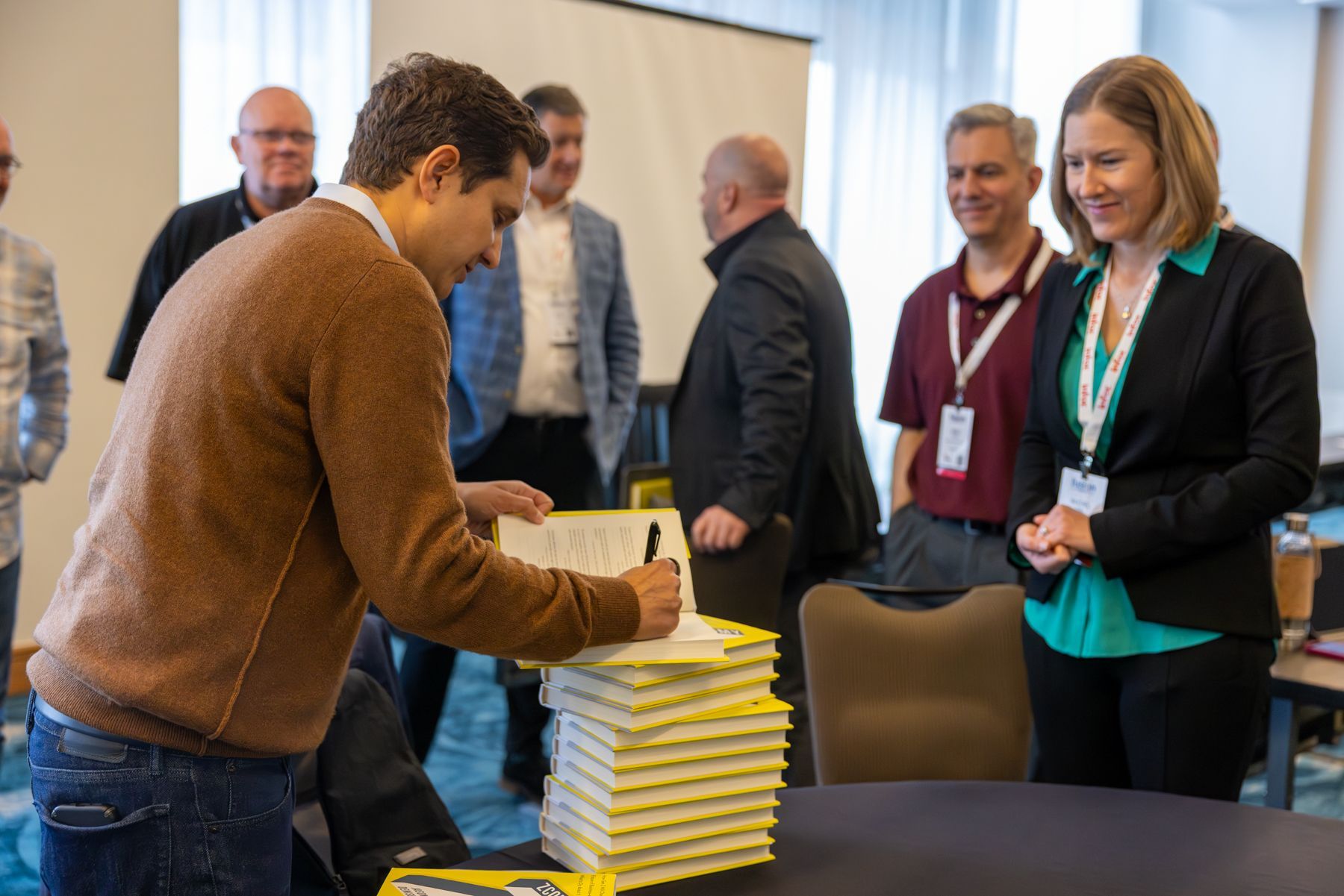 Man signing a stack of papers, observed by a woman and a group of people in a meeting setting.
