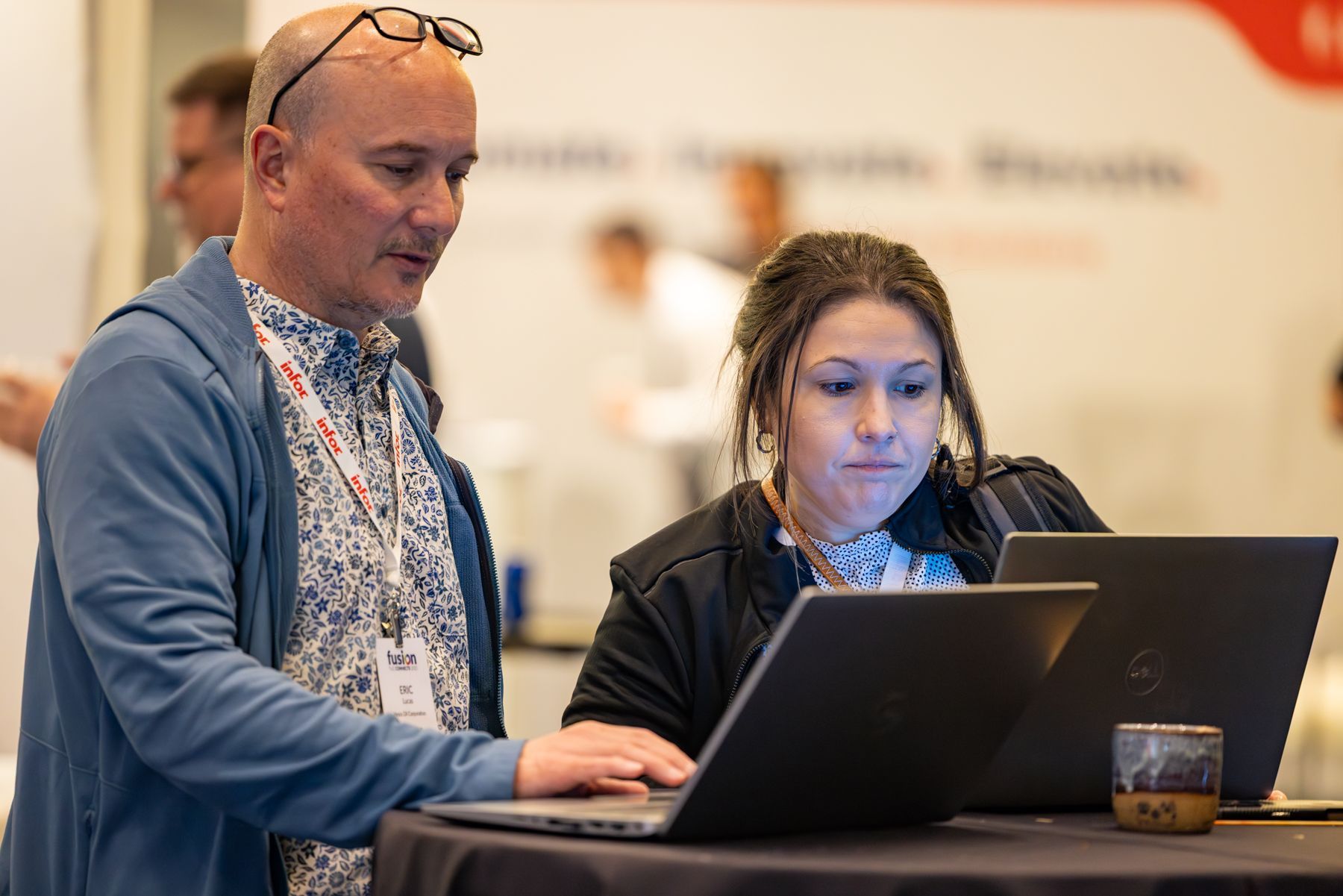 Two people at a table looking at laptop screens; man points, woman types. Indoors, blurred background.