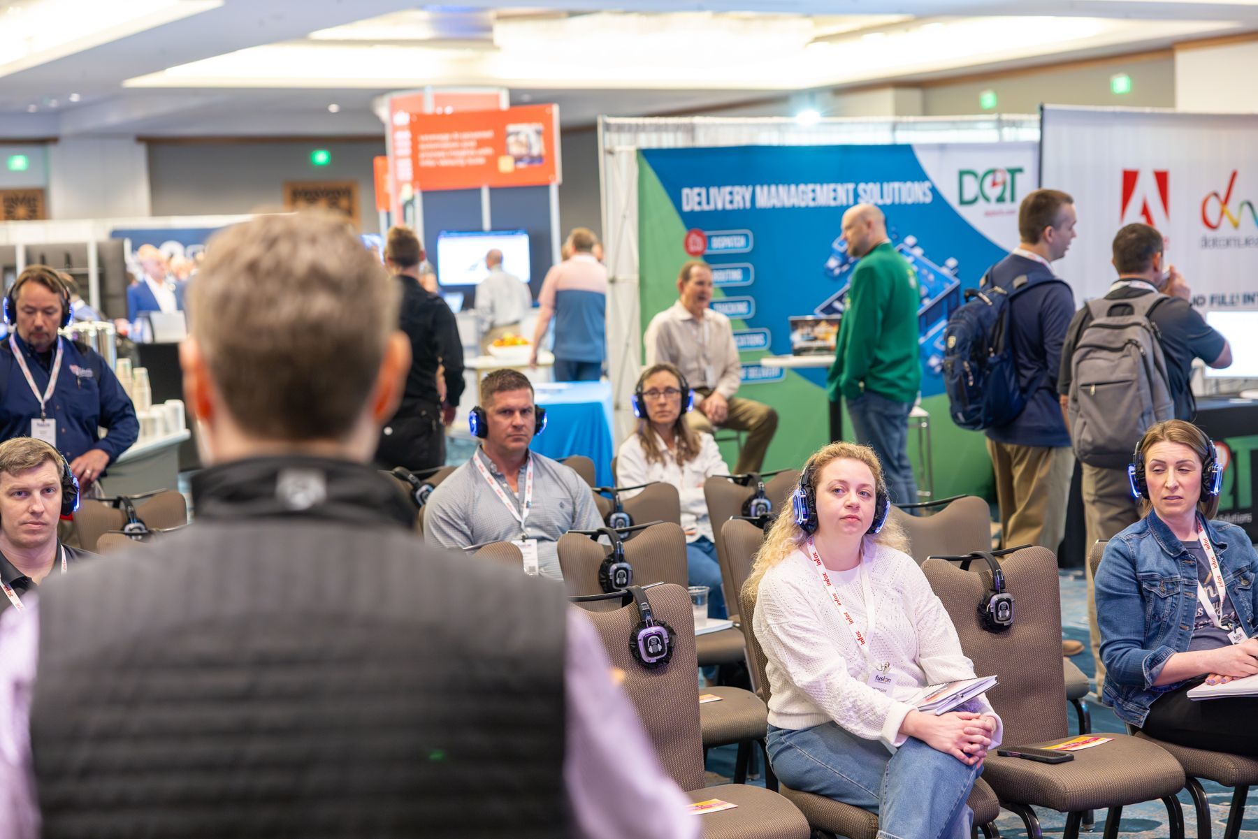 Audience with headphones at a conference. Some attendees look at a speaker; others stand near booths.