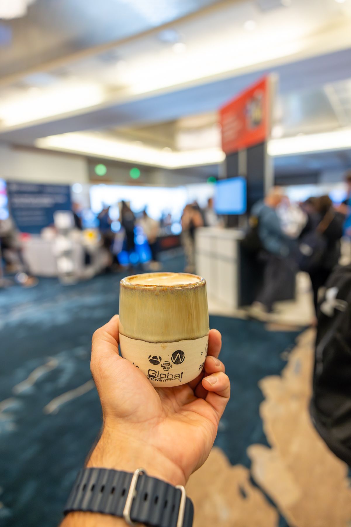 Hand holding a ceramic cup with tan top and brown pattern. Indoor setting, blurred background of a conference.