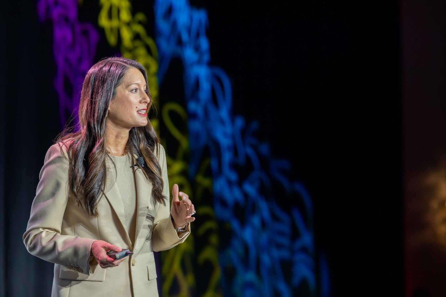 Woman in beige suit gestures while speaking on stage with colorful background.