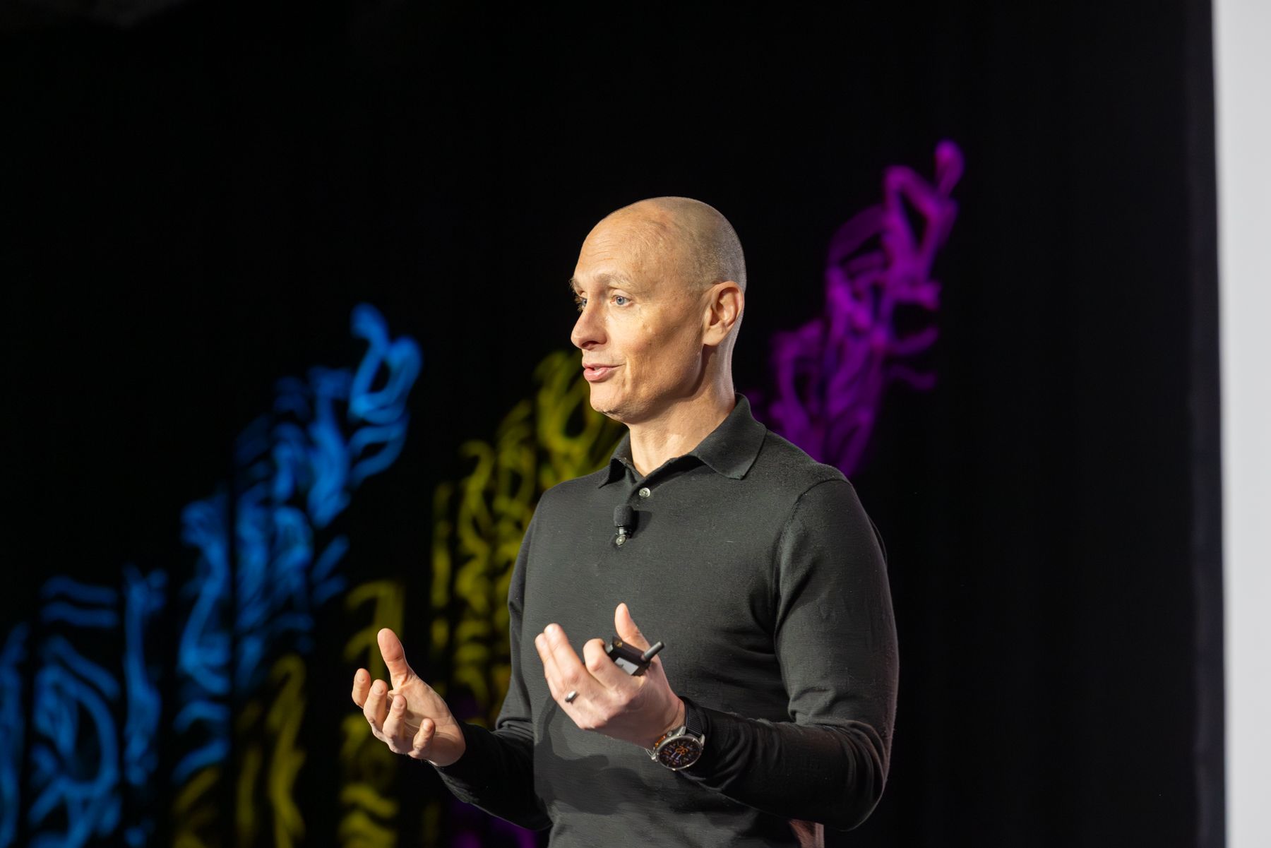 Man (Kevin Samuelson) with a shaved head speaking on stage with a colorful abstract background.