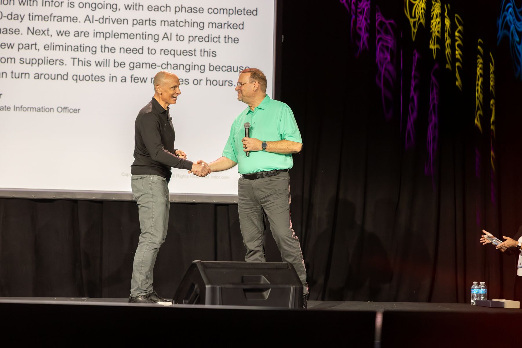 Two men shaking hands on a stage. One wears a gray shirt, the other a green polo. Text is visible behind them.