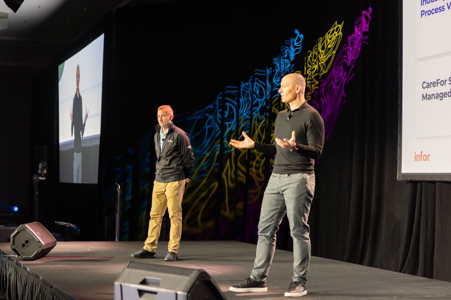 Two men on stage, one speaking with hands raised, other stands beside, large screen and abstract art backdrop.