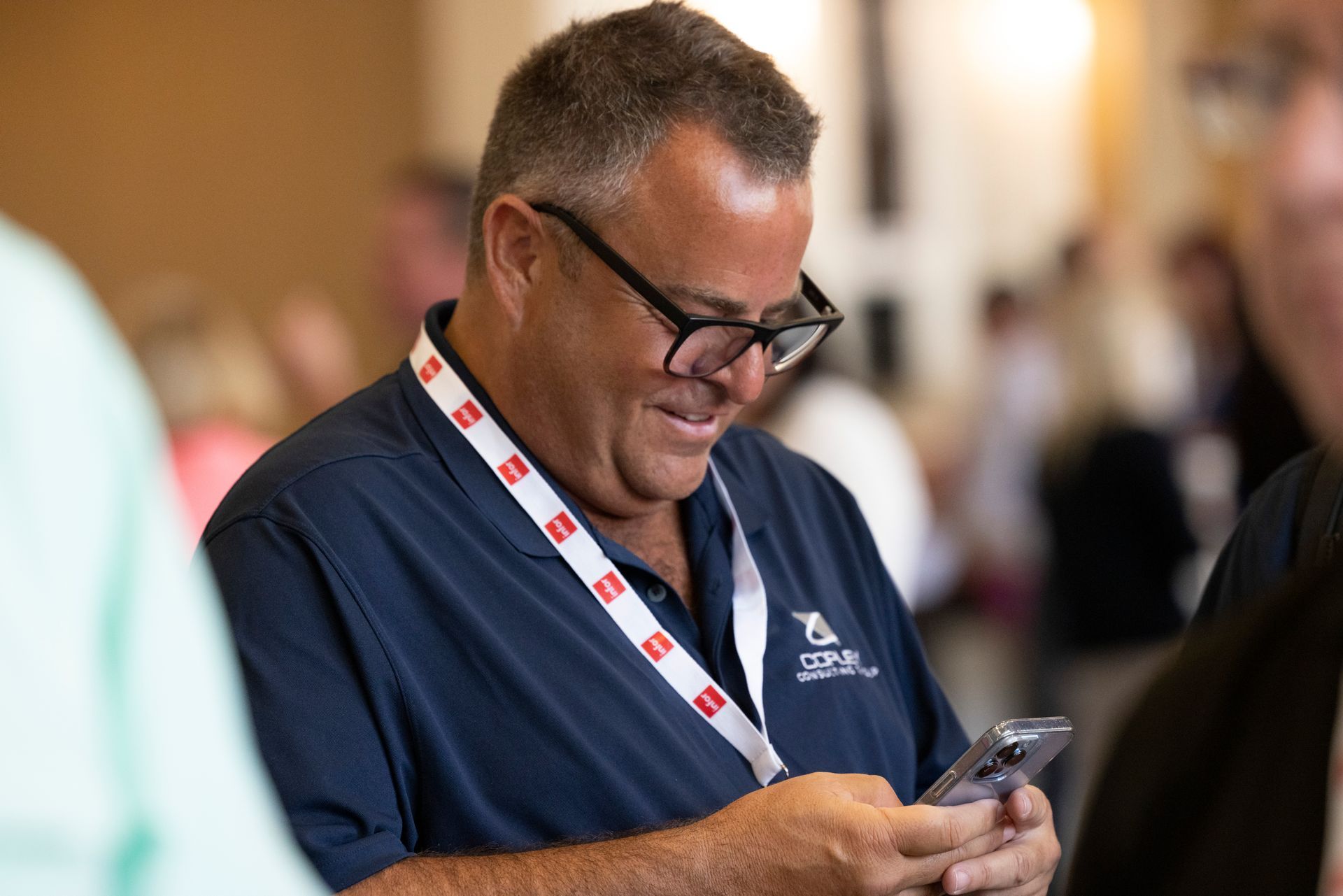 Man in glasses looks at phone, wearing a lanyard, smiling in a room with other people.