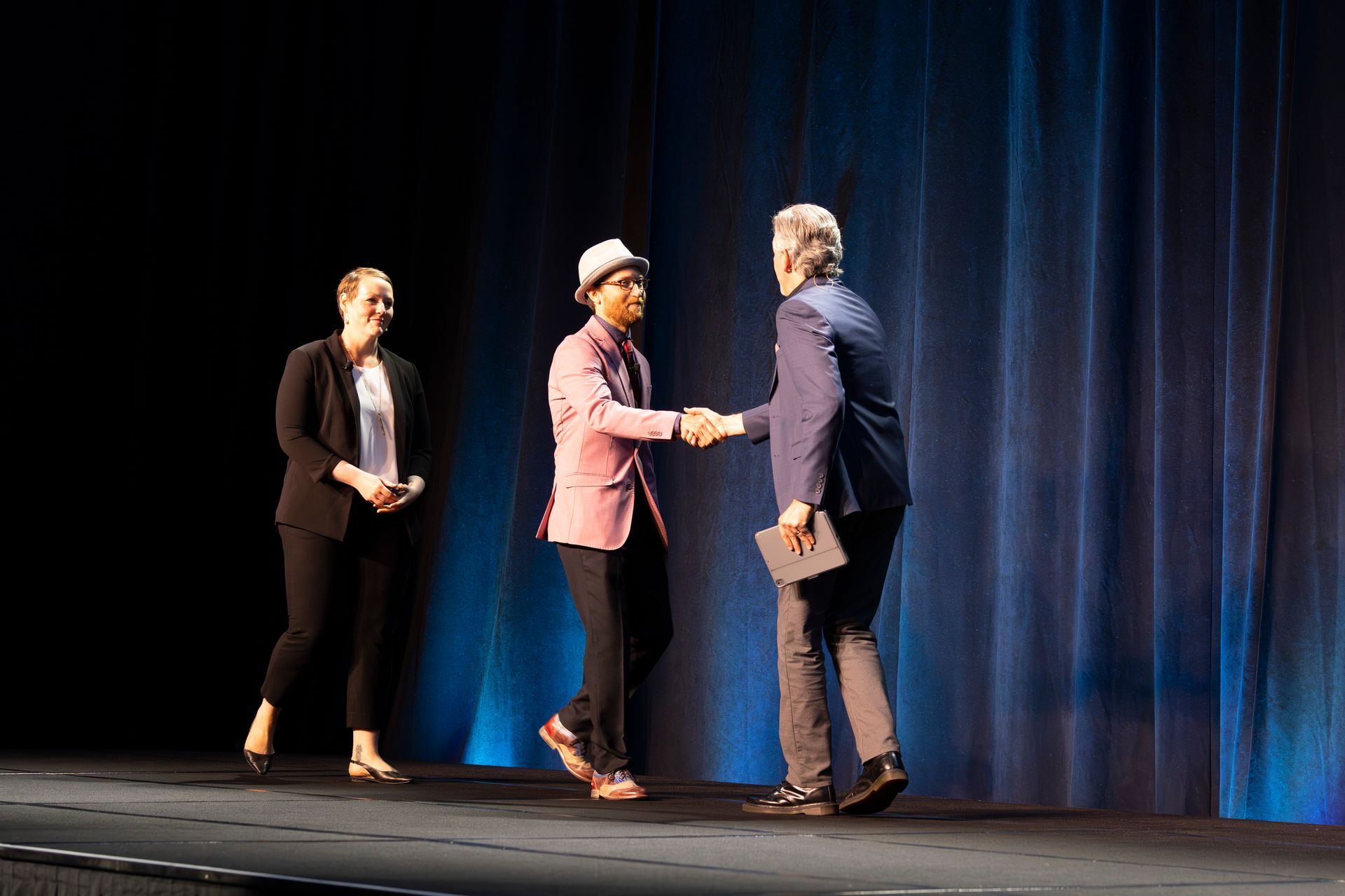 A person in a pink jacket shakes hands with another on a stage; a woman in a blazer stands by.