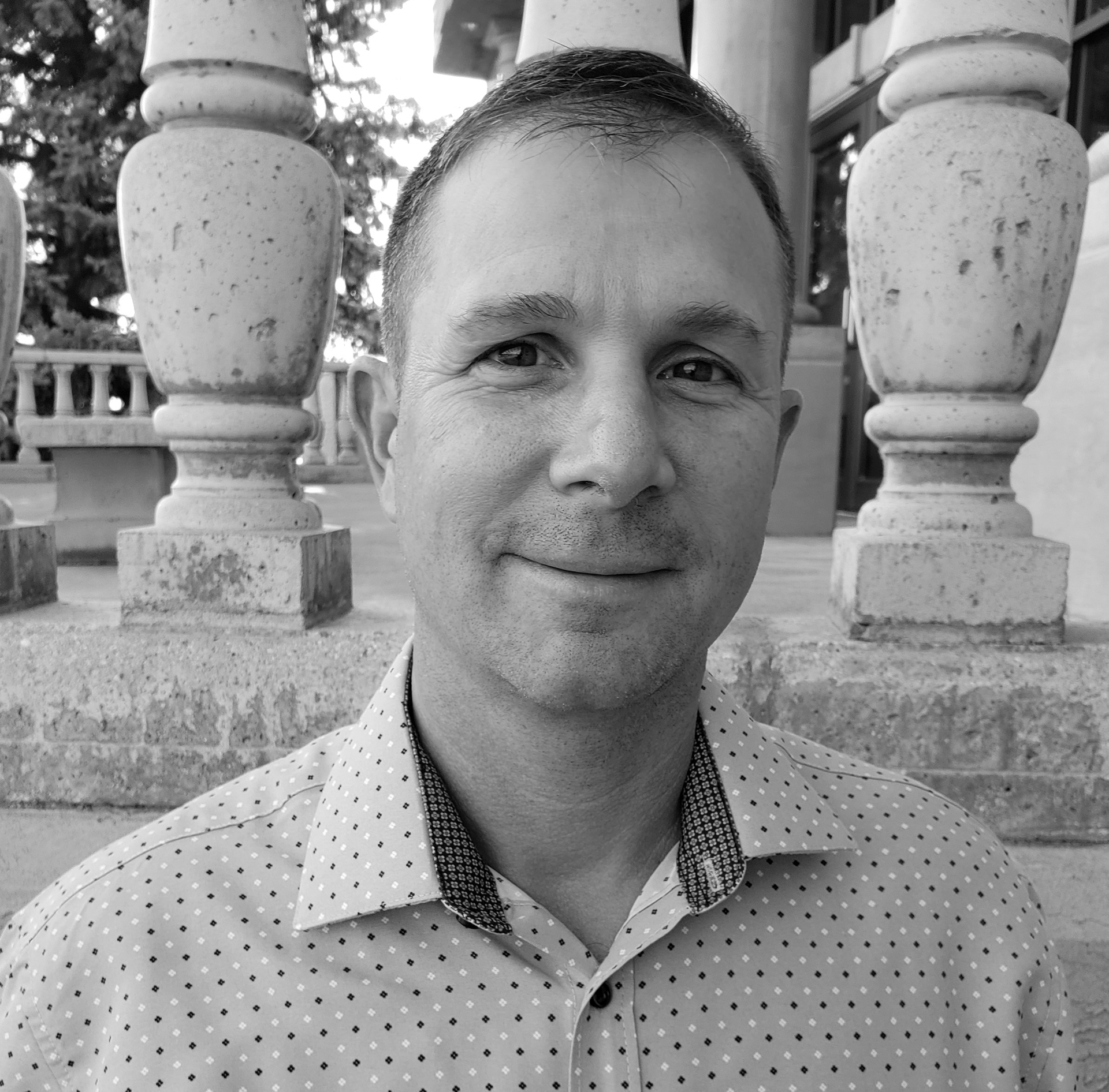 Man smiling in front of stone columns, wearing a collared patterned shirt.