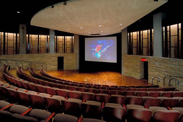 Auditorium with rows of maroon seats facing a screen. Stone walls surround the space.