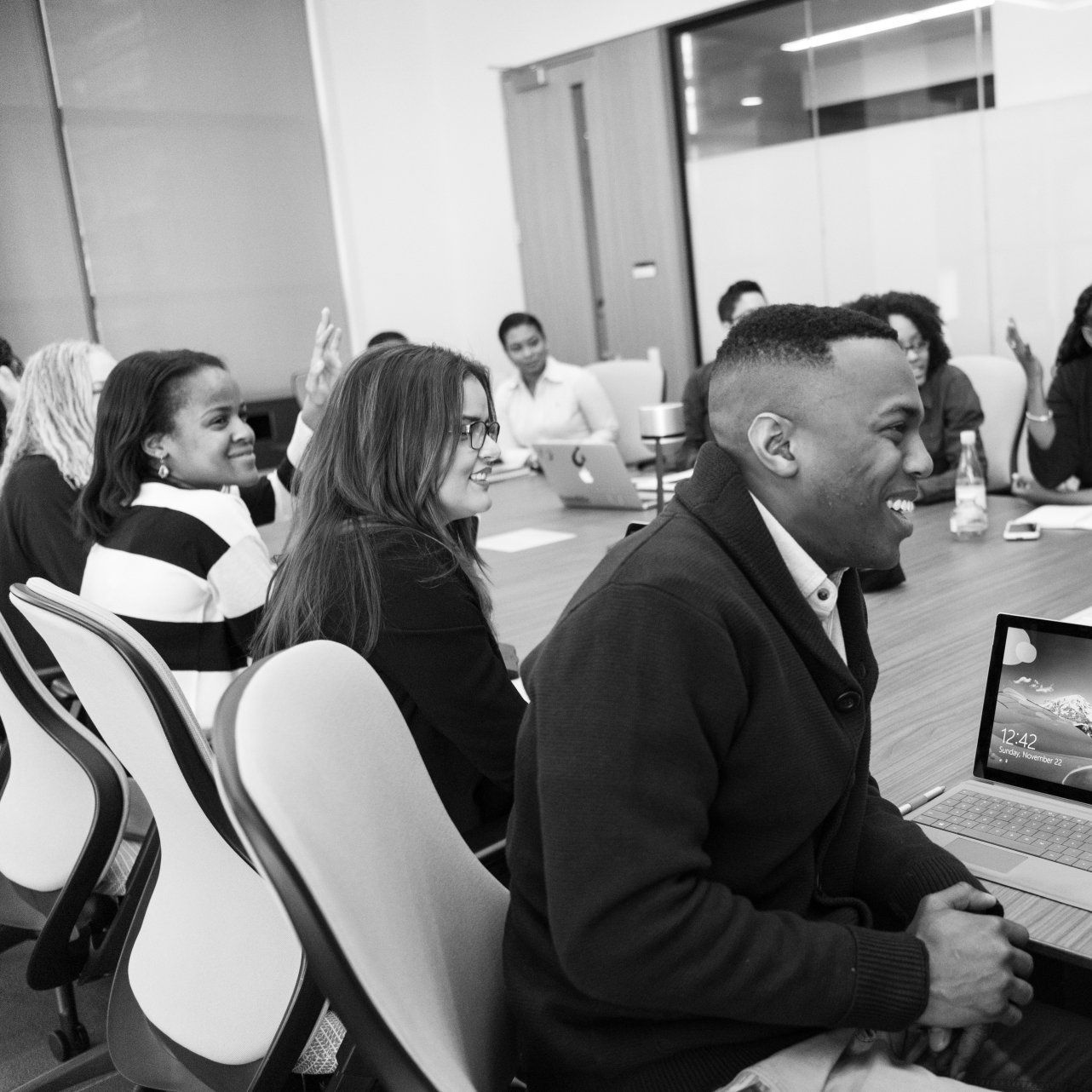 People in meeting around a table, some smiling, with laptops in a bright office space.