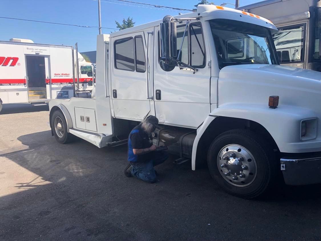 A mechanic repairs a white semi-truck in a paved lot on a sunny day.