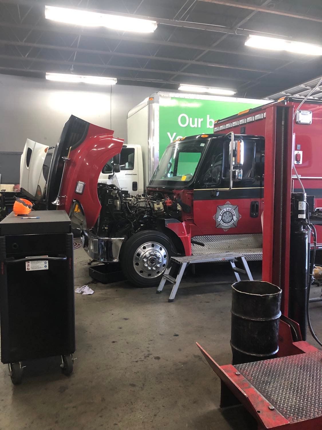Red fire truck in a repair shop with the hood open. Another truck in background.