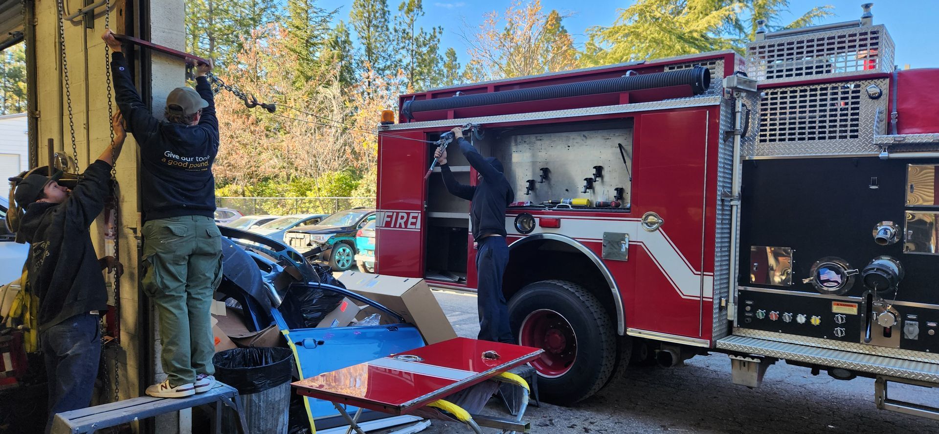 Firefighters working beside a red fire truck in a wooded area, handling equipment and a ladder. | Tripp's Auto Body