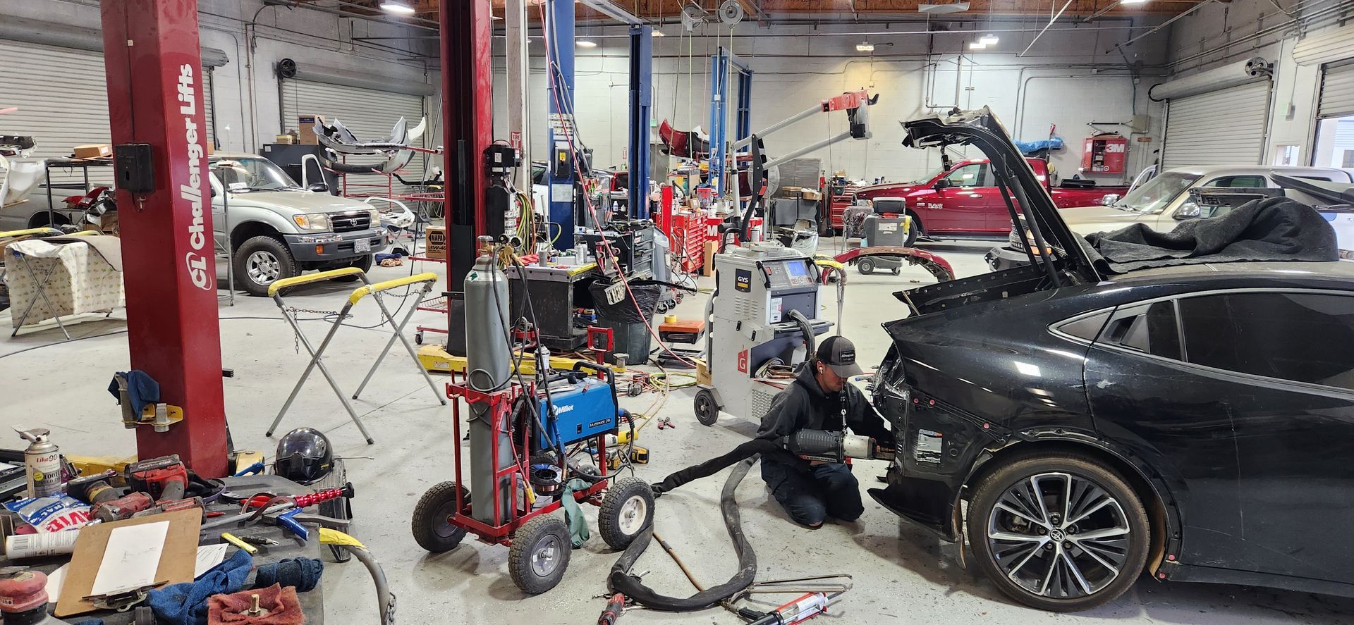 Auto repair shop with several cars, lifts, tools, and a worker servicing a black car on the right | Tripp's Auto Body