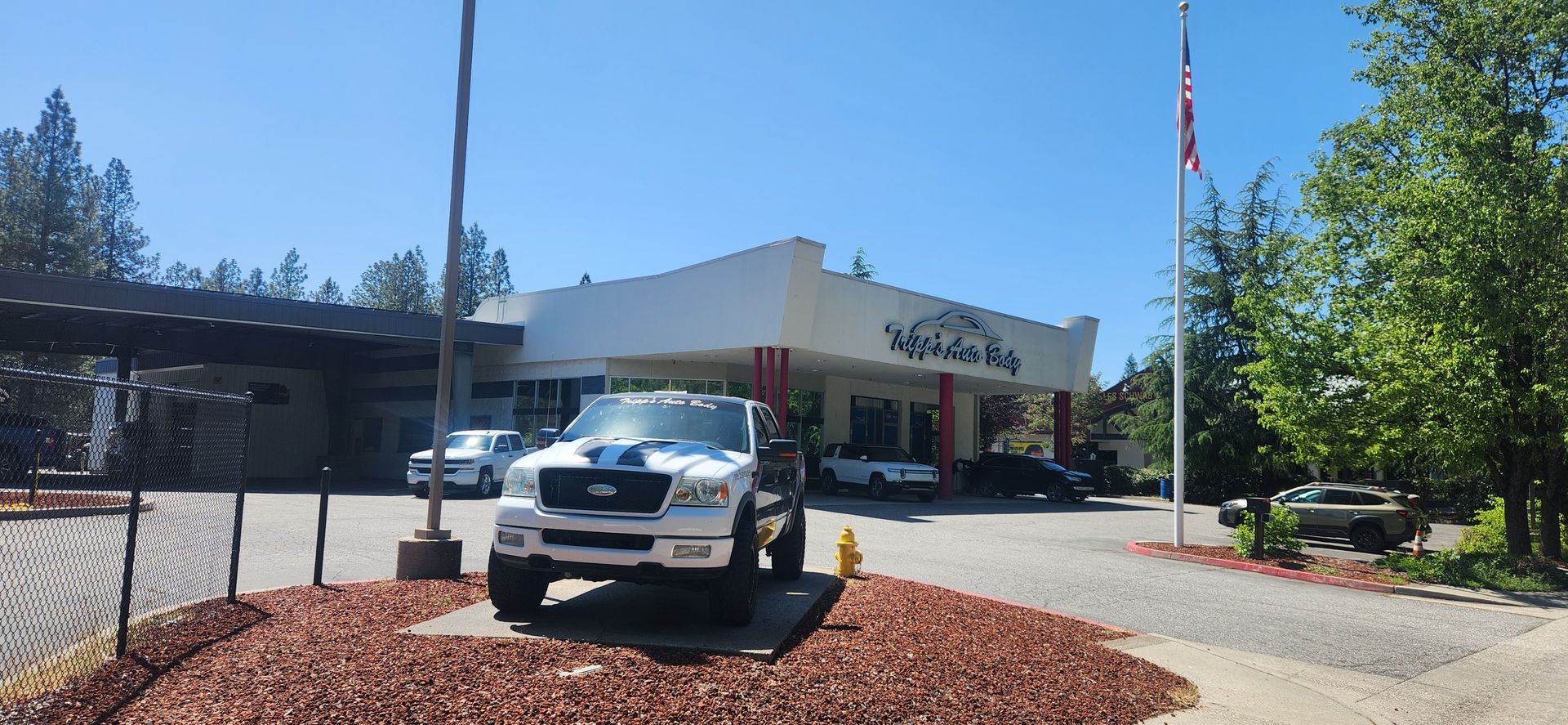 White pickup truck parked outside a building with an American flag and landscaped entrance | Tripp's Auto Body