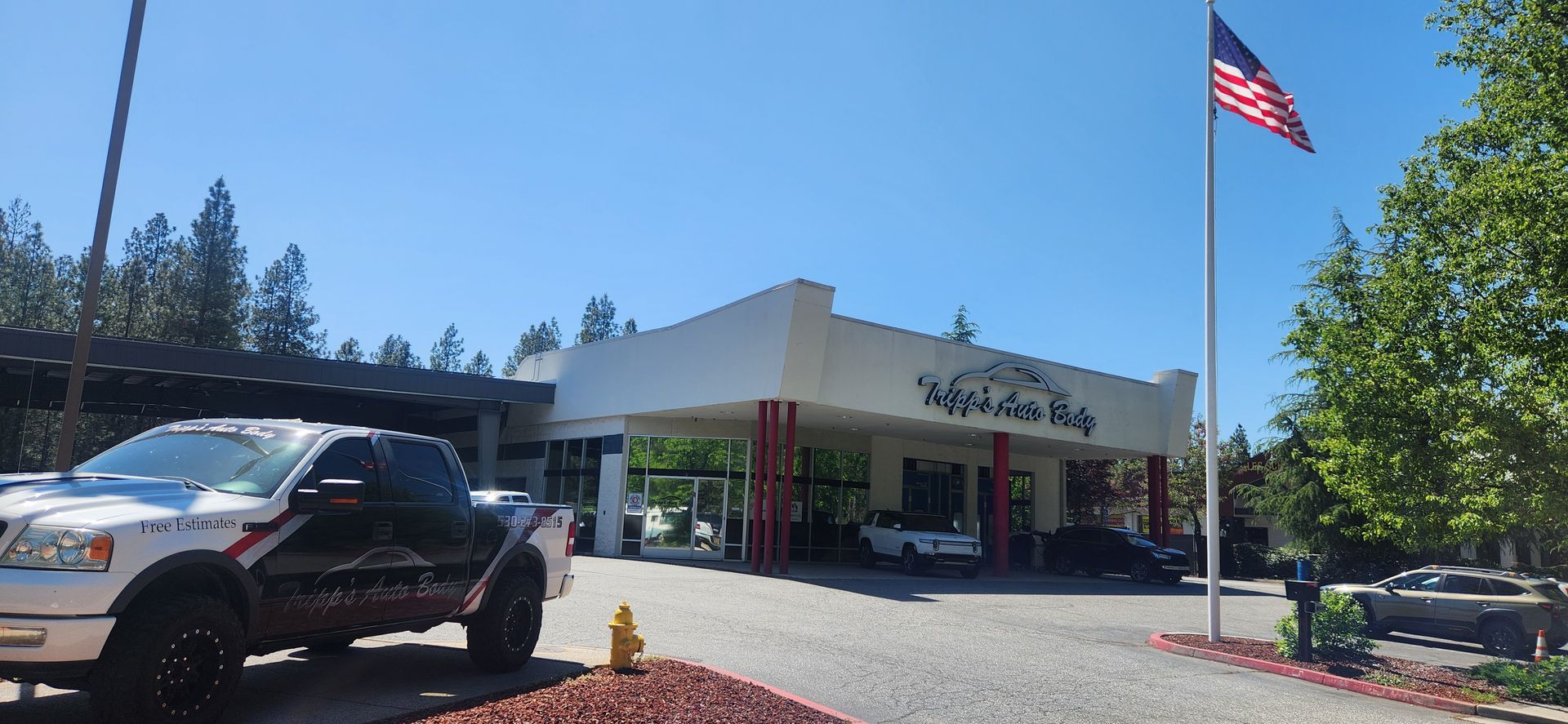 Auto Body shop exterior with parked cars and an American flag on a sunny day | Tripp's Auto Body