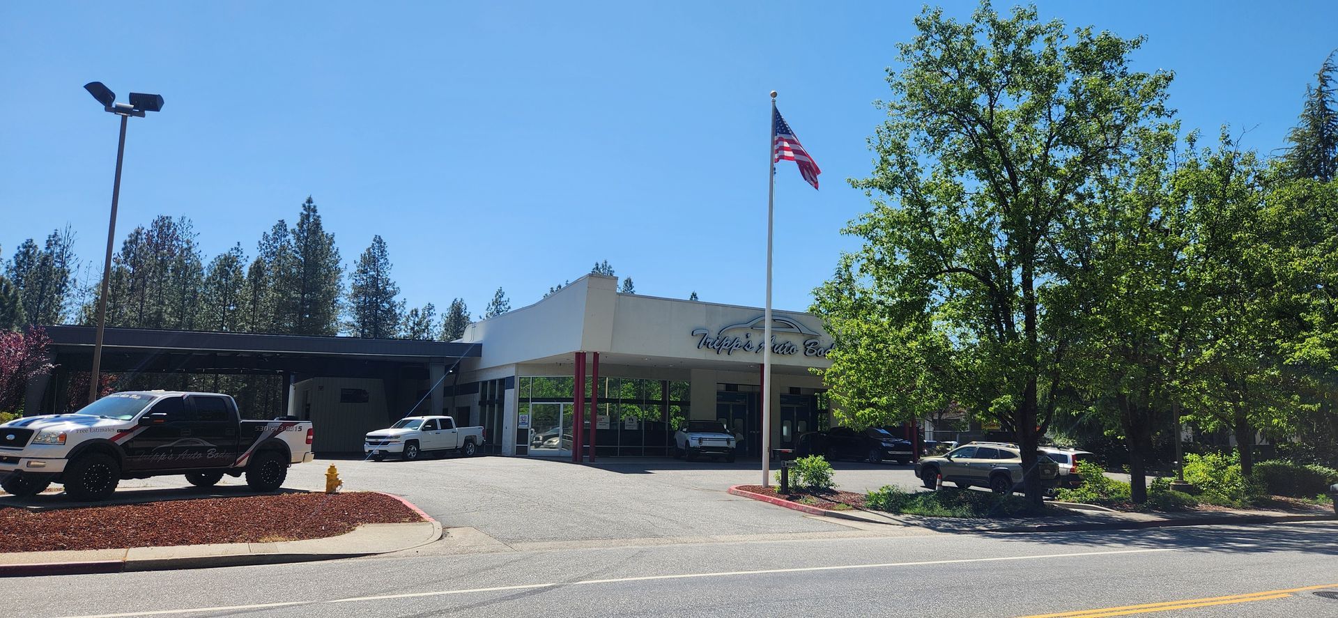 Low building entrance with American flag, parked vehicles, and trees on a sunny day | Tripp's Auto Body