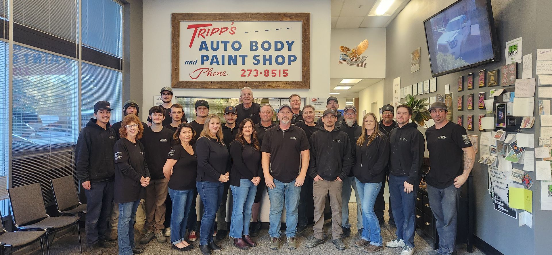 Group of staff posing in front of a “Auto Body & Paint Shop” sign inside a workshop | Tripp's Auto Body